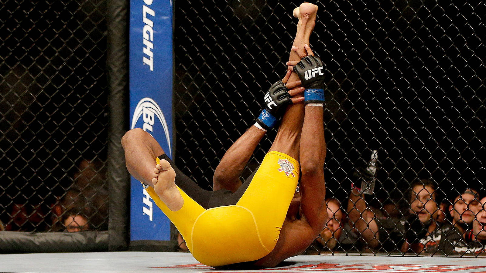 UFC 桌布 壁紙 — 一名身穿黃色短褲的拳手在UFC八角籠內對對手施展腿鎖，觀眾正在觀看。