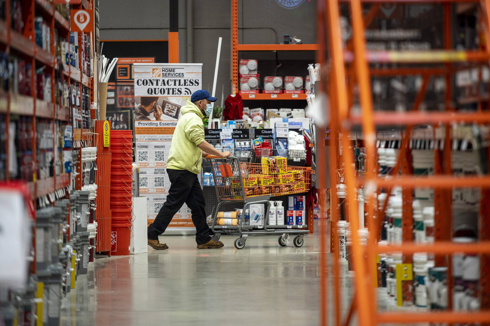 Home Depot 桌布 壁紙 — 一名身穿黃色背心的人推著裝滿貨物的手推車，穿過光線充足、擺放橙色貨架的倉庫通道。