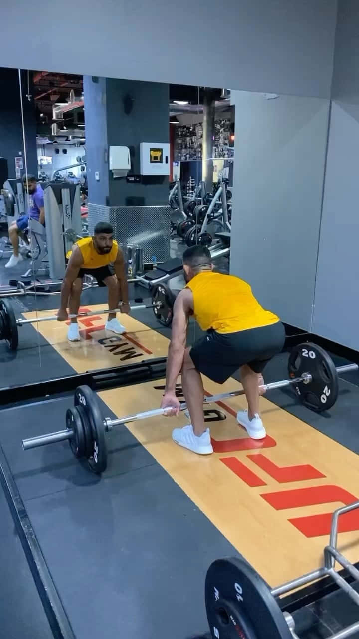 Fitness 桌布 壁紙 — 一名身穿黃色背心的男子在現代健身房中進行槓鈴硬舉，動作反映在鏡中。