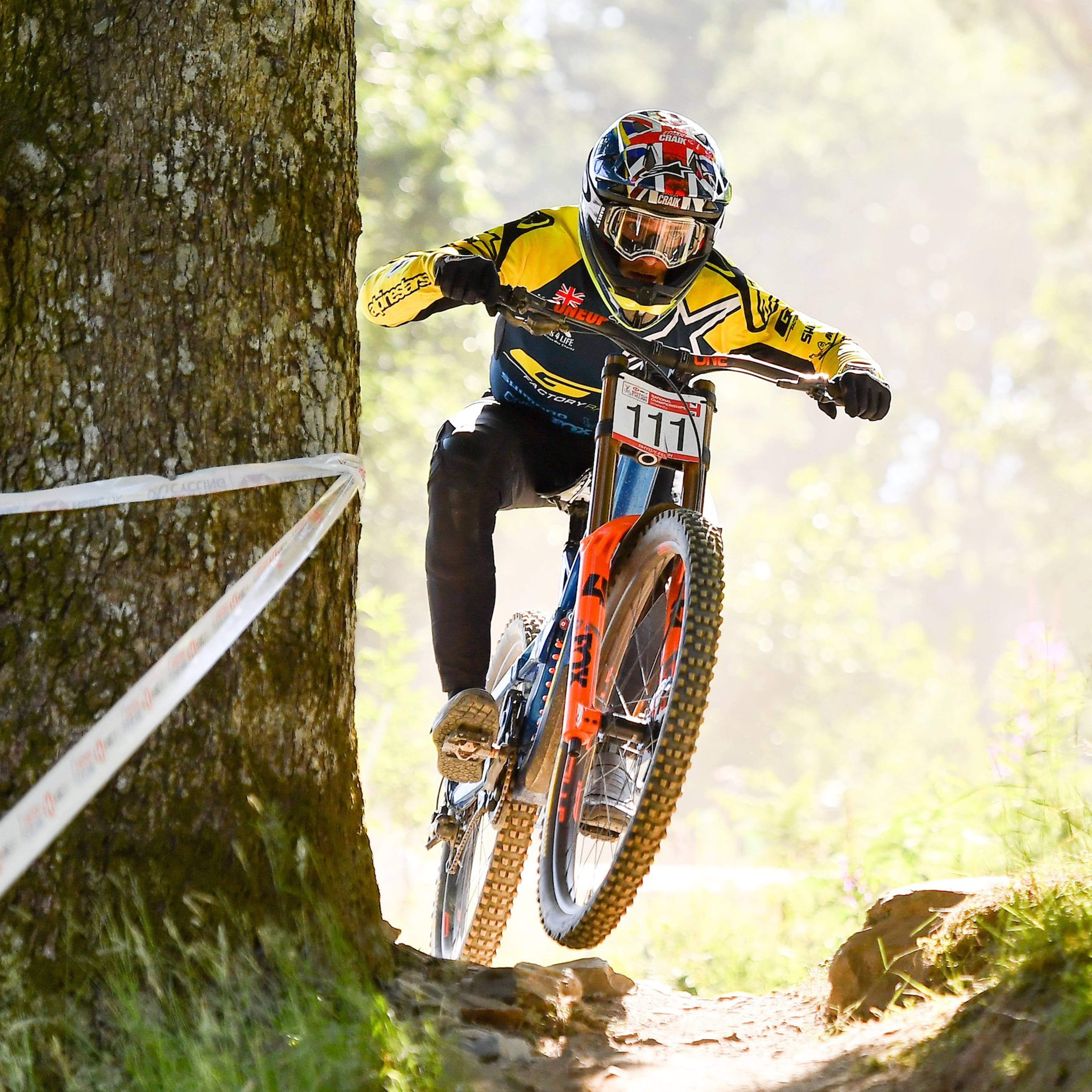 Mountain Bike 桌布 壁紙 — 一名身穿黃色裝備的山地自行車手在陽光穿過樹林的森林小徑上騰空跳躍。