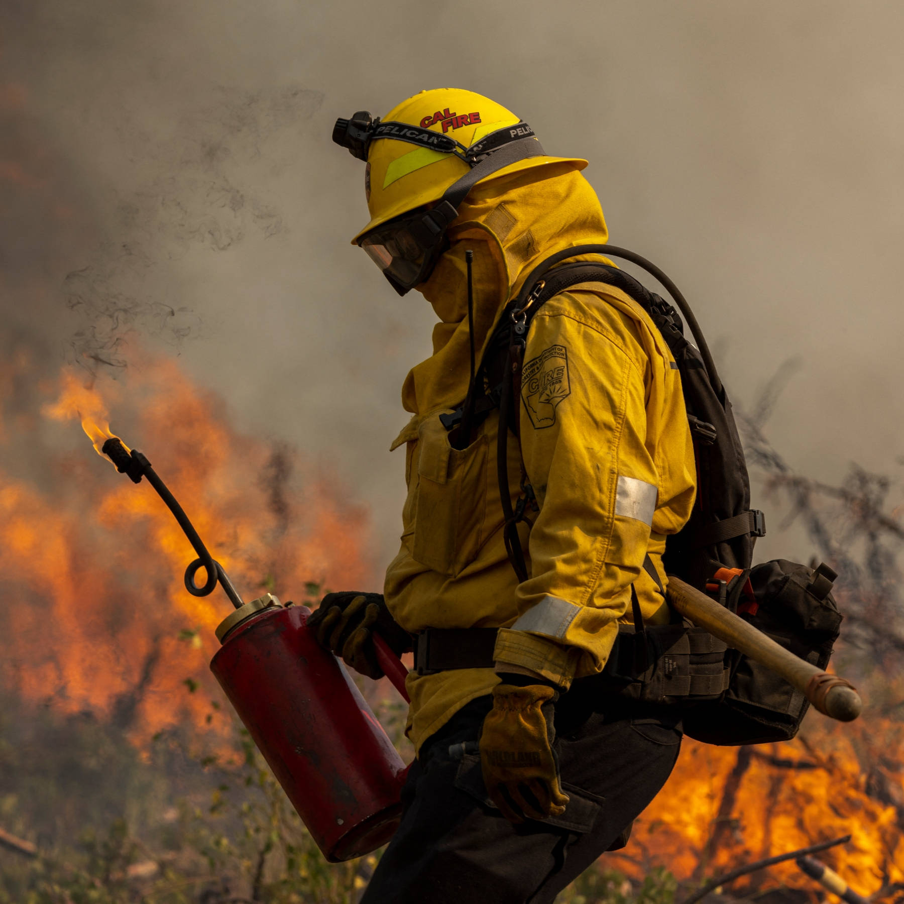 Firefighter 桌布 壁紙 — 一名身穿黃色裝備的消防員在煙霧中前進，手持燃燒的火把，背景是野火。