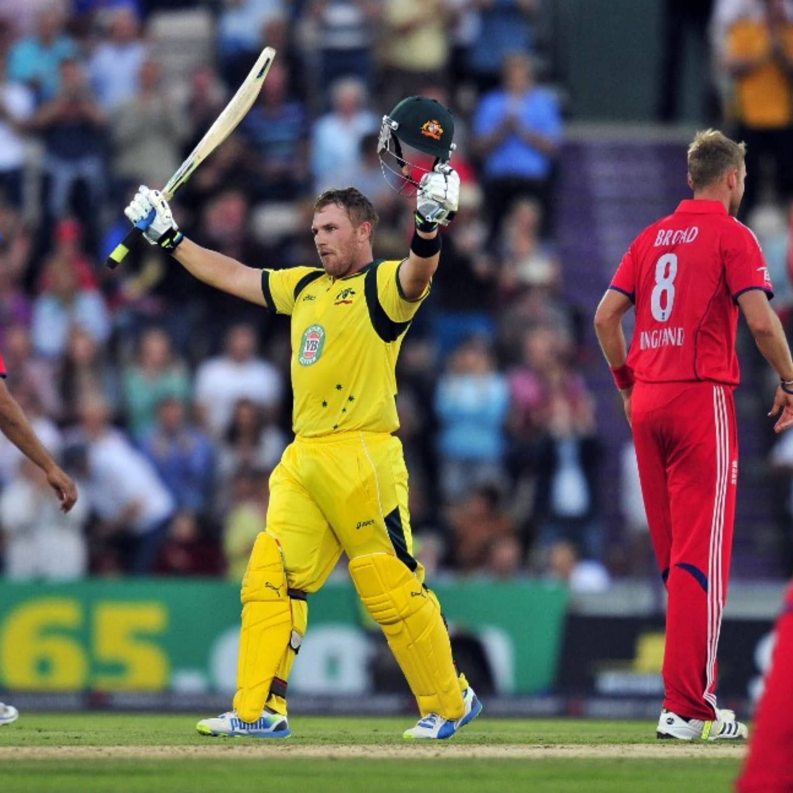 Aaron Finch 桌布 壁紙 — 一名身穿黃色裝備的澳洲板球運動員在綠色球場上，背景是模糊的人群，勝利地高舉球棒和頭盔。