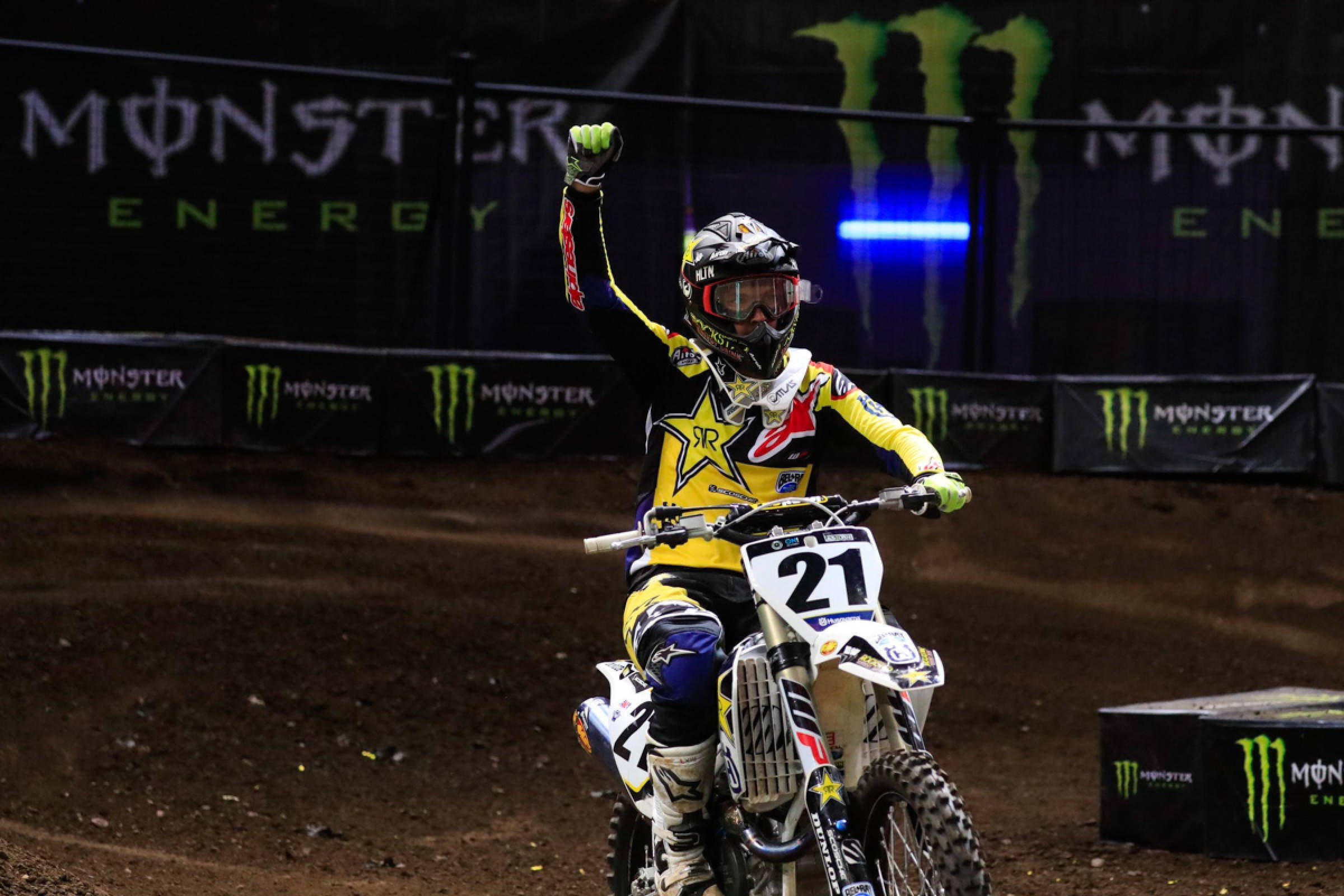 Ronnie Mac 桌布 壁紙 — 一名身穿黃色裝備的越野摩托車手，在明亮的場館燈光下，於泥地賽道上慶祝勝利，並帶有Monster Energy品牌標誌。