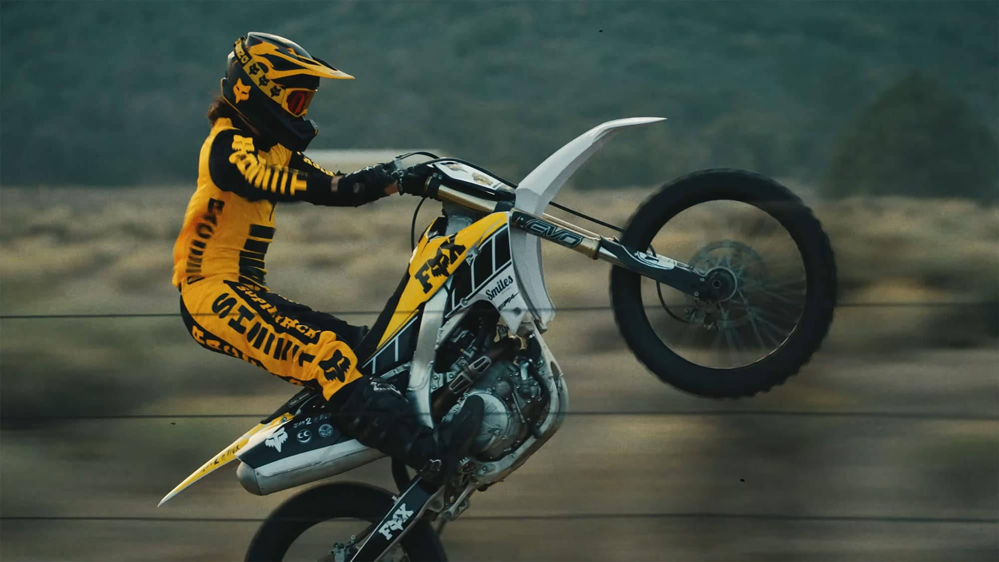 Motocross 桌布 壁紙 — 一名身穿黃色裝備的越野摩托車手在模糊的自然背景下表演獨輪行駛。