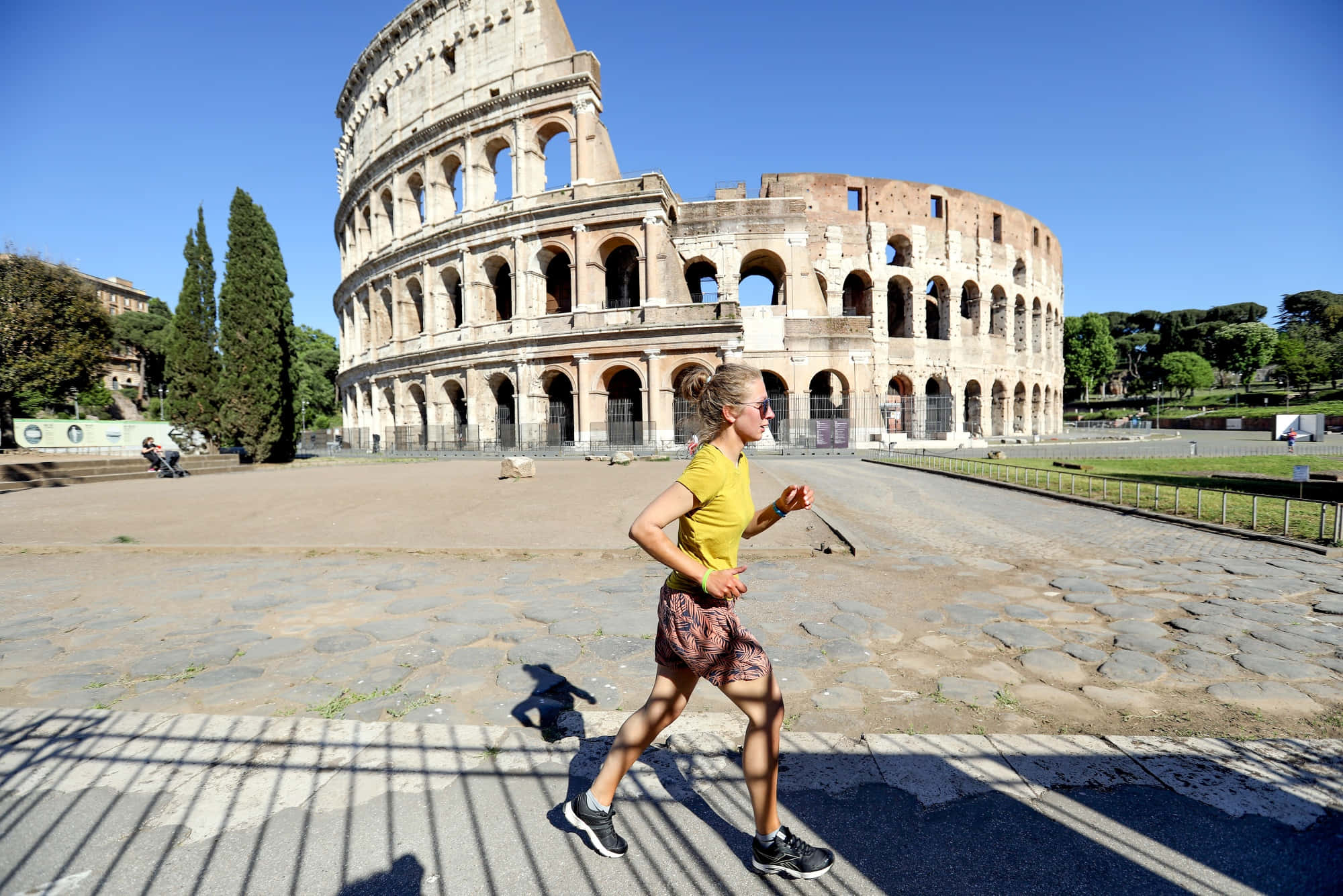 Roman Colosseum 桌布 壁紙 — 一名身穿黃色襯衫的女子在明亮的藍天下，帶著強烈的陰影跑過古羅馬競技場。