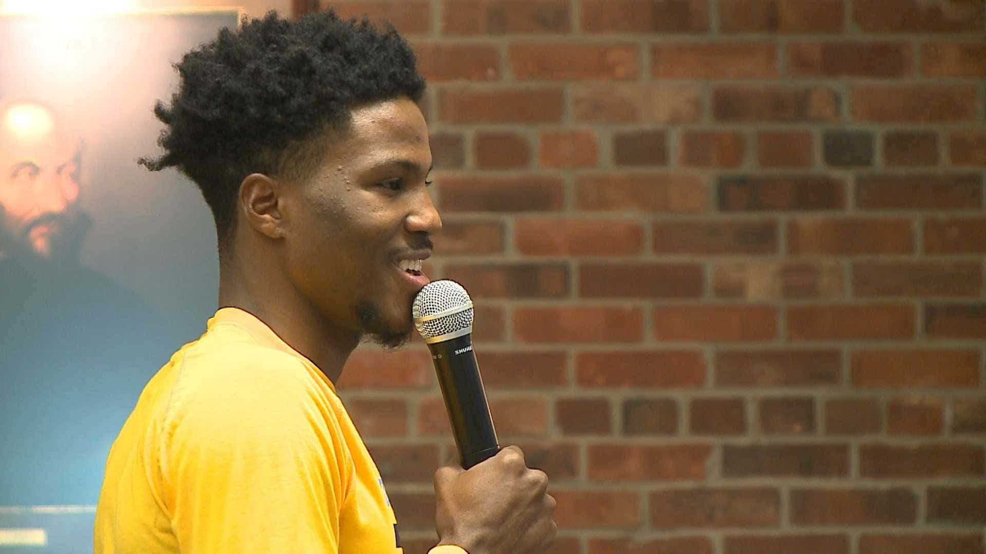 籃球 桌布 壁紙 — 一名身穿黃色襯衫的年輕男子對著麥克風說話，背景是紅磚牆和一幅海報。