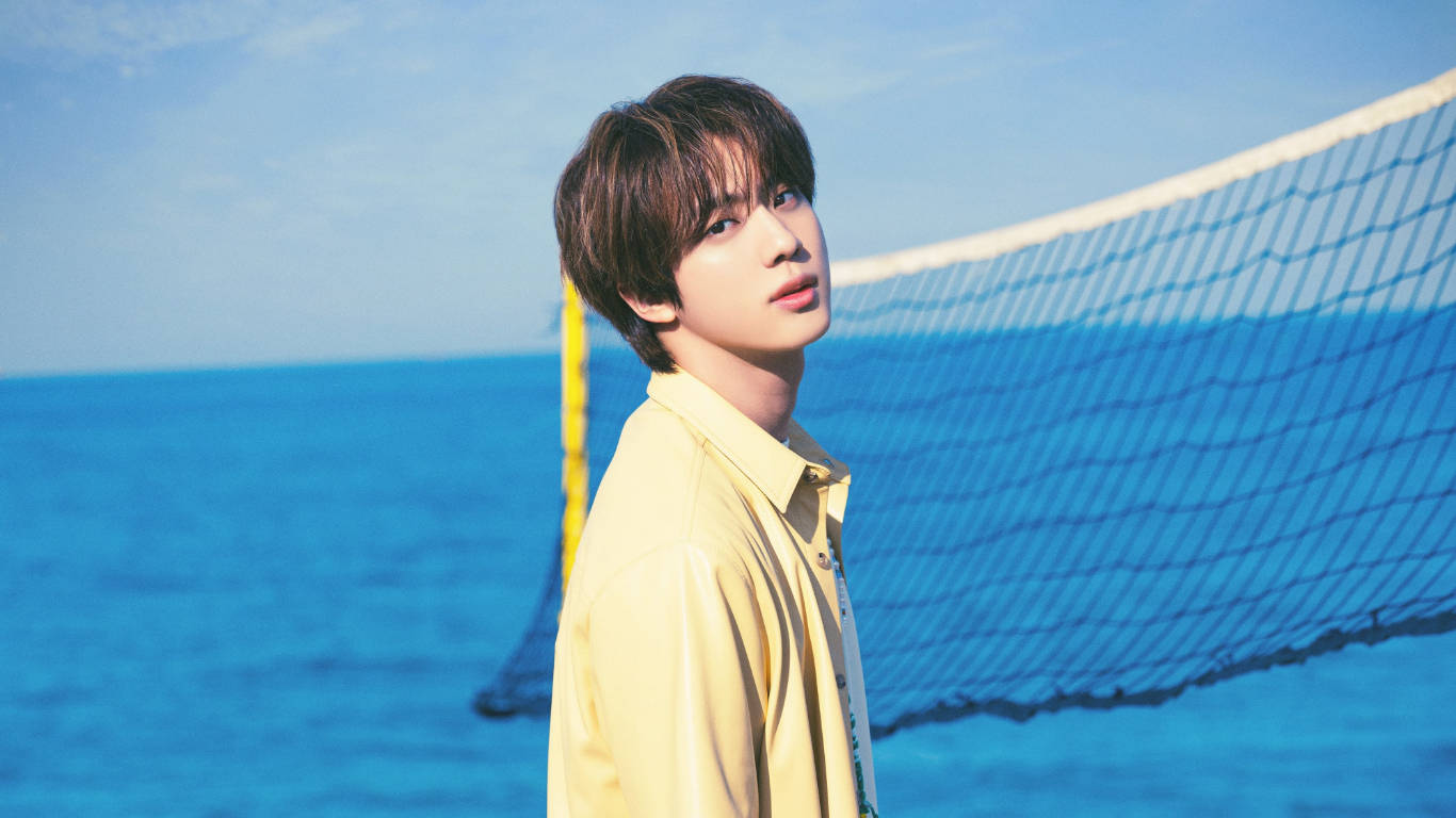 kim seok jin 桌布 壁紙 — 一名身穿黃色襯衫的年輕男子站在排球網旁邊，背景是明亮的藍色海洋和天空。