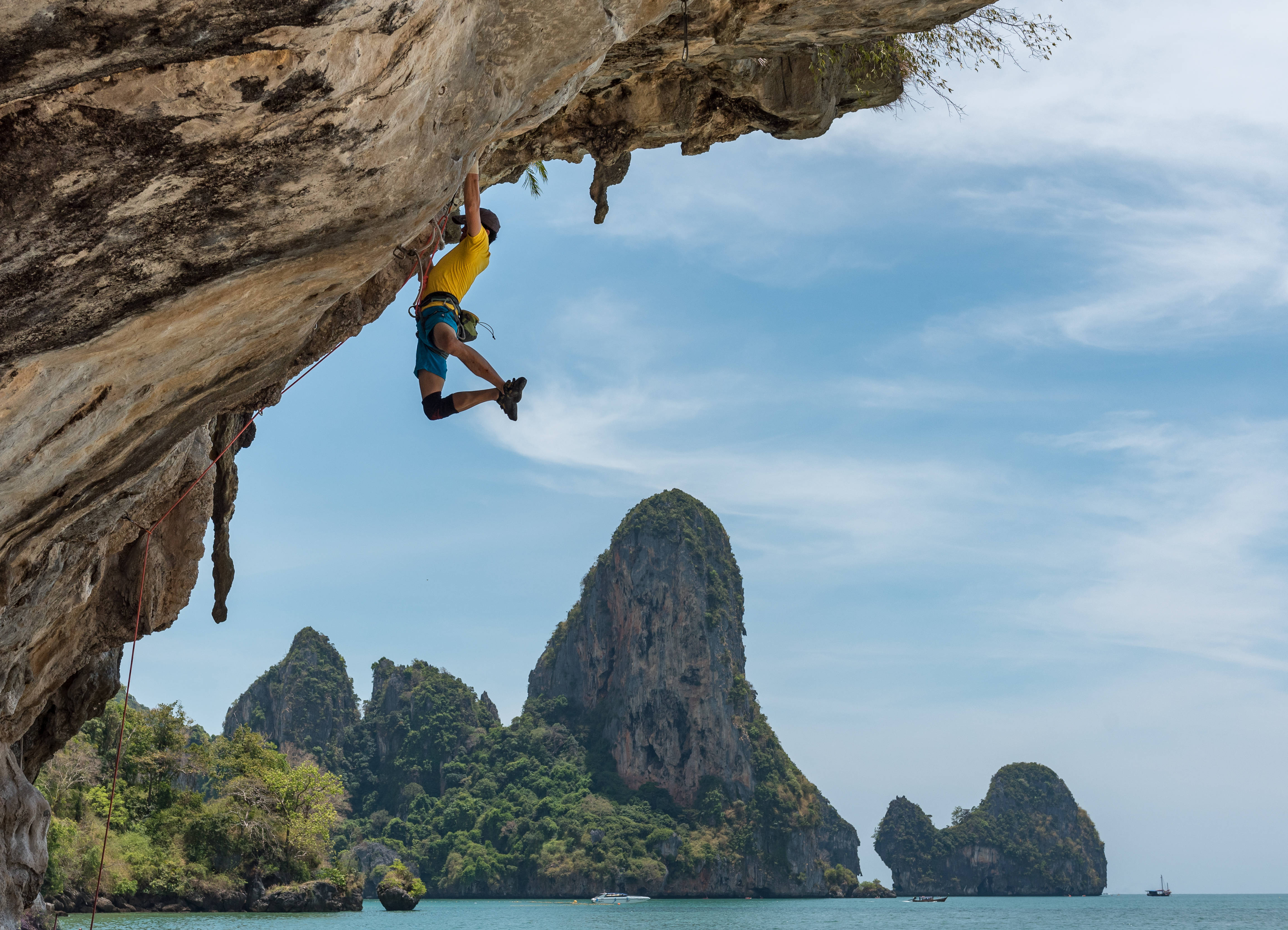 Climbing 桌布 壁紙 — 一名身穿黃色襯衫的攀岩者正在攀爬石灰岩懸崖，下方是碧綠色的海水，遠處是叢林覆蓋的喀斯特地貌，天空湛藍明亮。