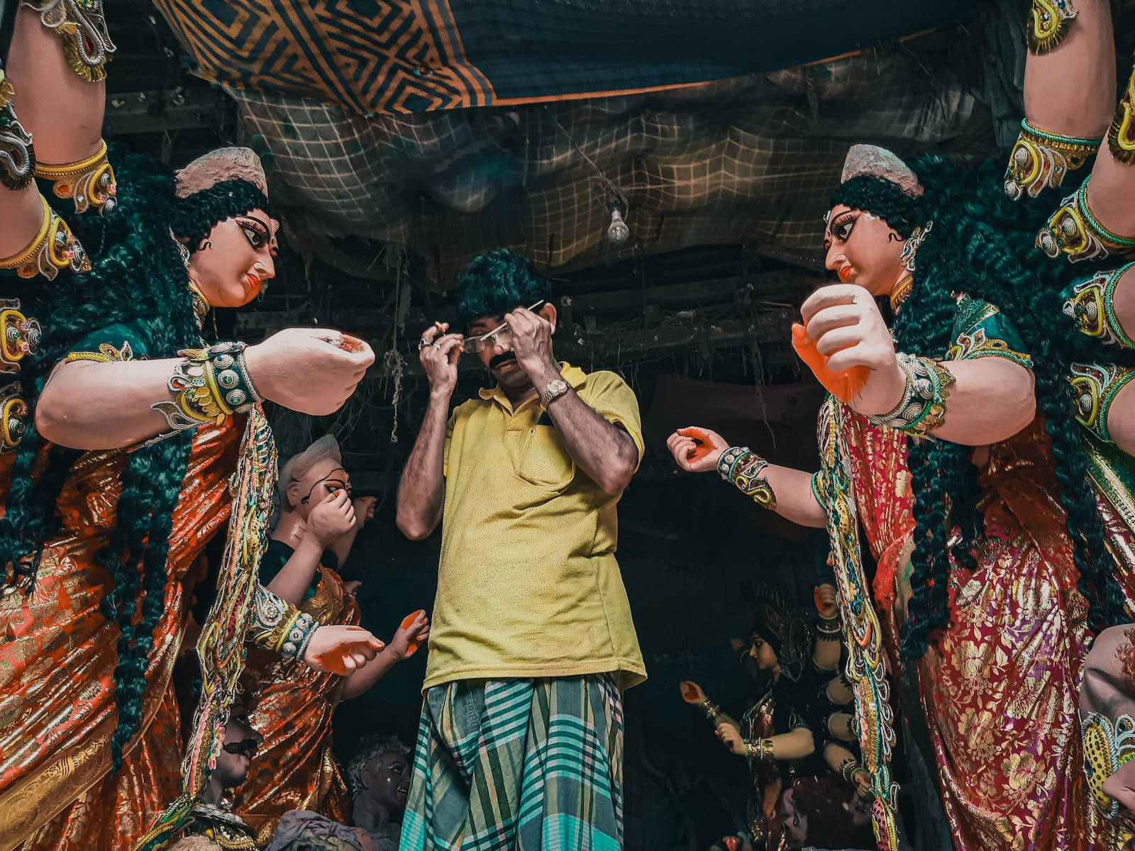 maa durga 桌布 壁紙 — 一名身穿黃色襯衫的男子在光線昏暗的寺廟中，被高聳華麗的杜爾迦雕像包圍，正在調整眼鏡。
