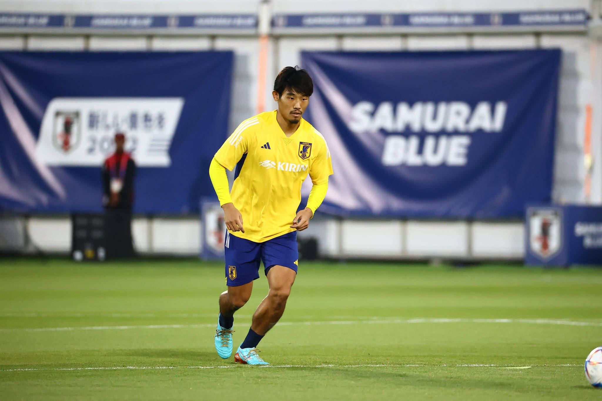 Football Players 桌布 壁紙 — 一名身穿黃色訓練服的日本足球員在綠色球場上奔跑，身後有 'SAMURAI BLUE' 的橫幅。