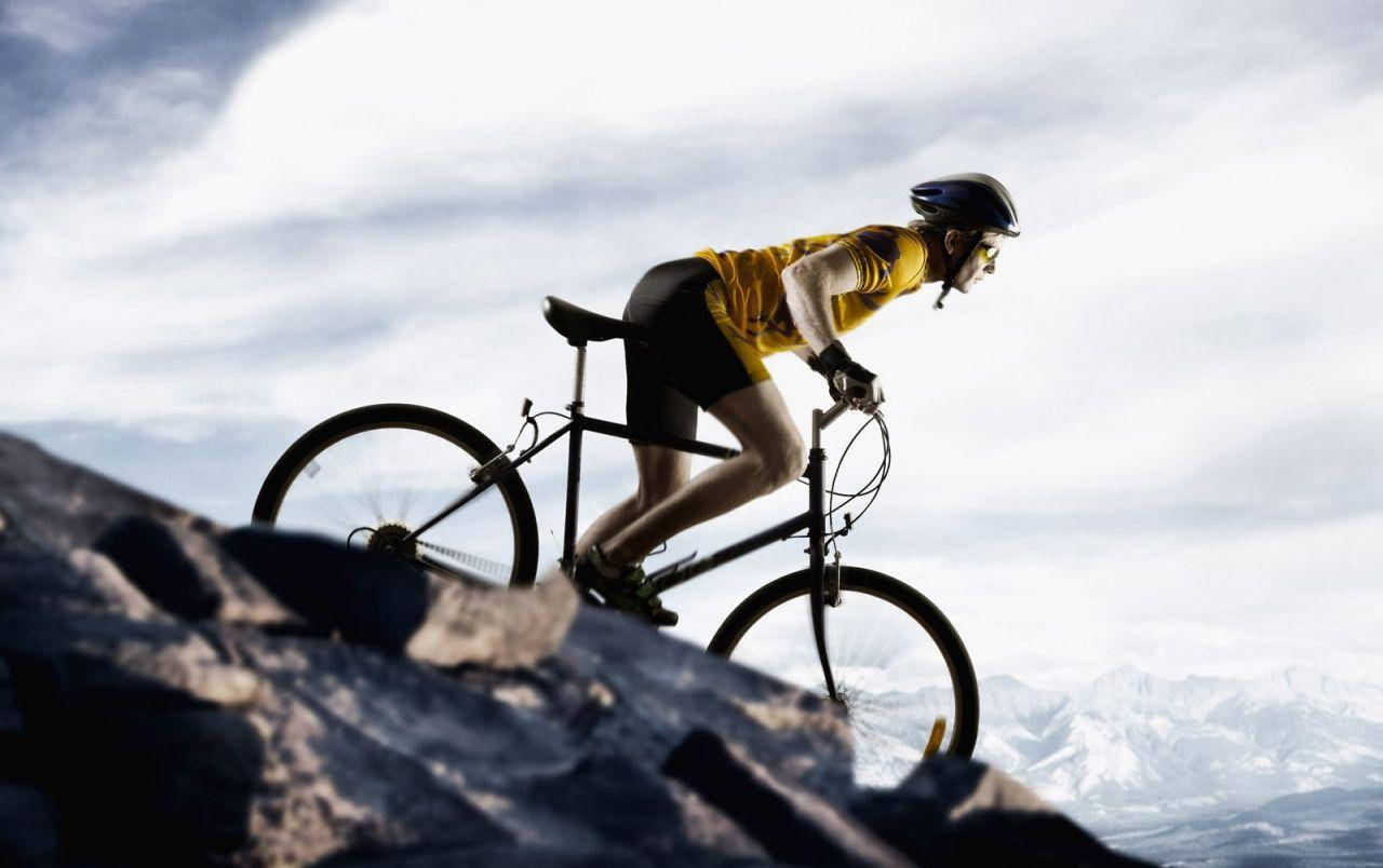 bike riding 桌布 壁紙 — 一名身穿黃色車衣的自行車手在岩石小徑上踩踏而上，背景是雲天下積雪覆蓋的山峰。