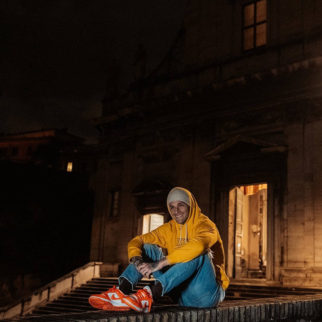 Athlete 桌布 壁紙 — 一名身穿黃色連帽衫和橙色運動鞋的年輕男子在夜間坐在燈火通明的歷史建築前的石階上微笑。