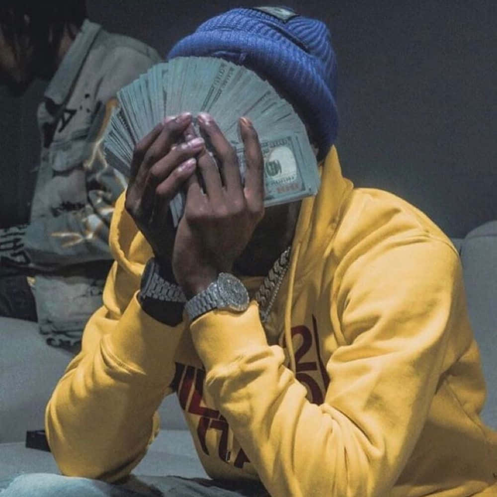 Youngboy 桌布 壁紙 — 一名身穿黃色連帽衫和藍色無邊帽的人坐在室內，手持一疊現金遮住臉部。