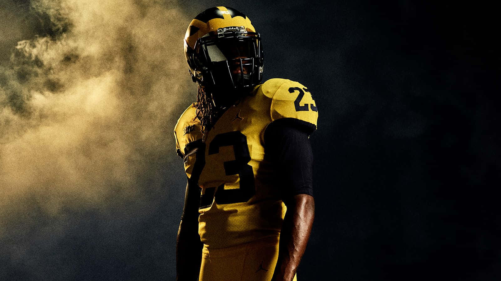 Michigan Wolverines 桌布 壁紙 — 一名身穿黃黑球衣的足球員在黑暗的攝影棚中，以金色煙霧為背景形成剪影。