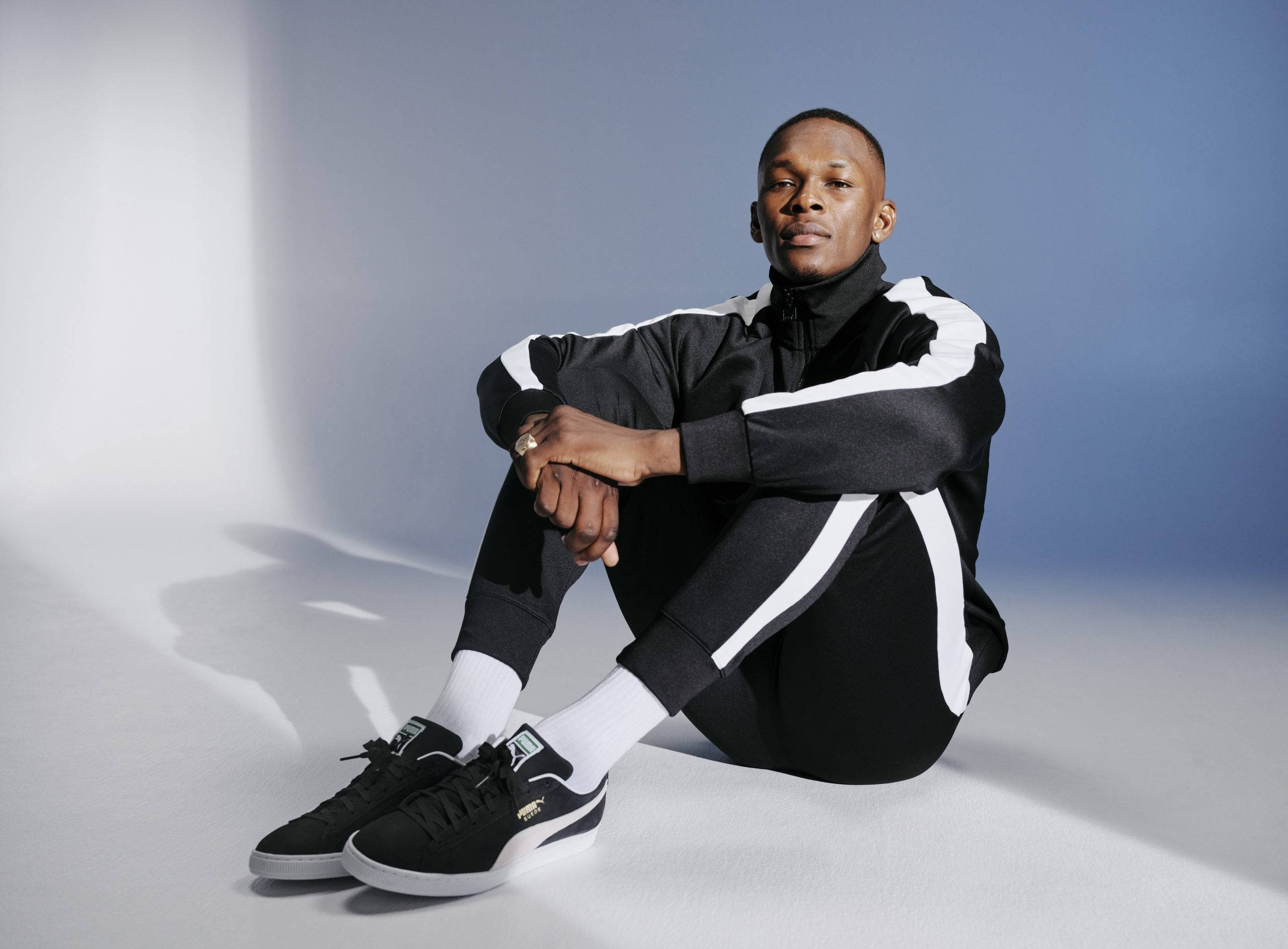 Adesanya 桌布 壁紙 — 一名身穿黑白運動服的男人自信地坐在工作室地板上，背景是藍白漸變色。