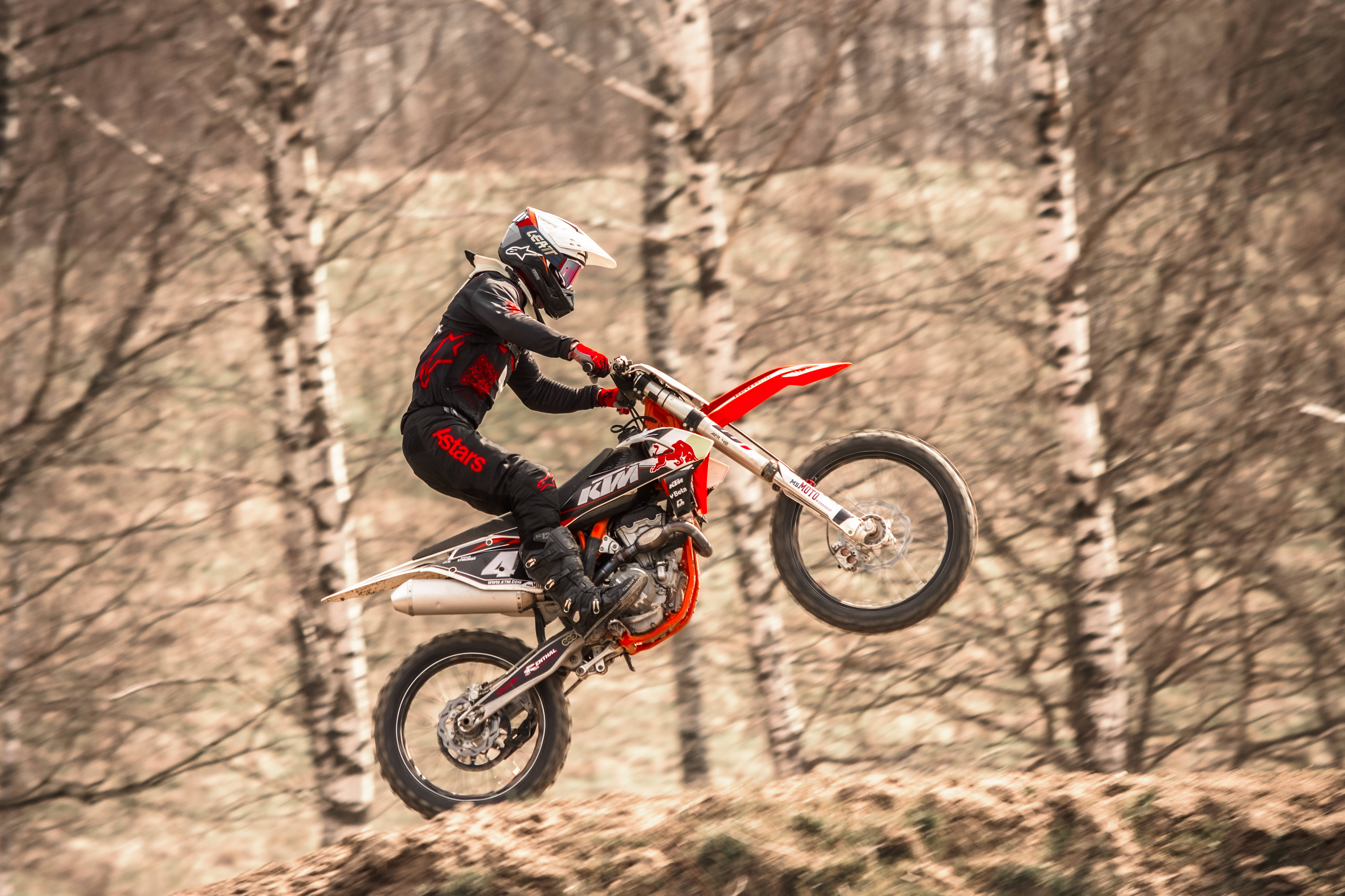 Ktm Bike 桌布 壁紙 — 一名身穿黑紅色裝備的越野機車手，騎著紅色KTM機車，在光禿禿的白樺樹環繞的泥徑上空飛翔。