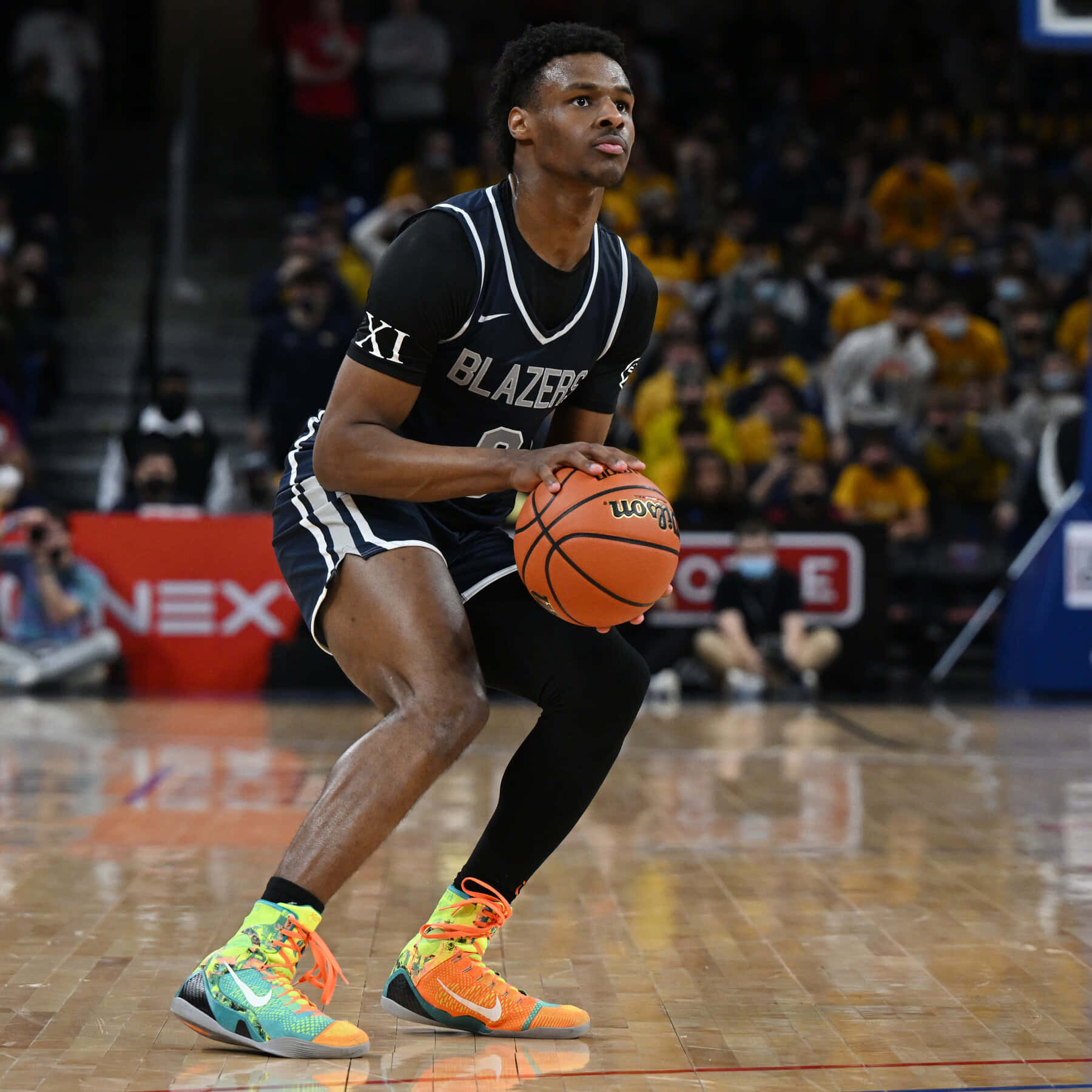 Bronny James 桌布 壁紙 — 一名身穿黑色 'Blazers' 球衣的籃球運動員在拋光球場上運著橙色籃球，身後有穿著黃色球衣的模糊觀眾。