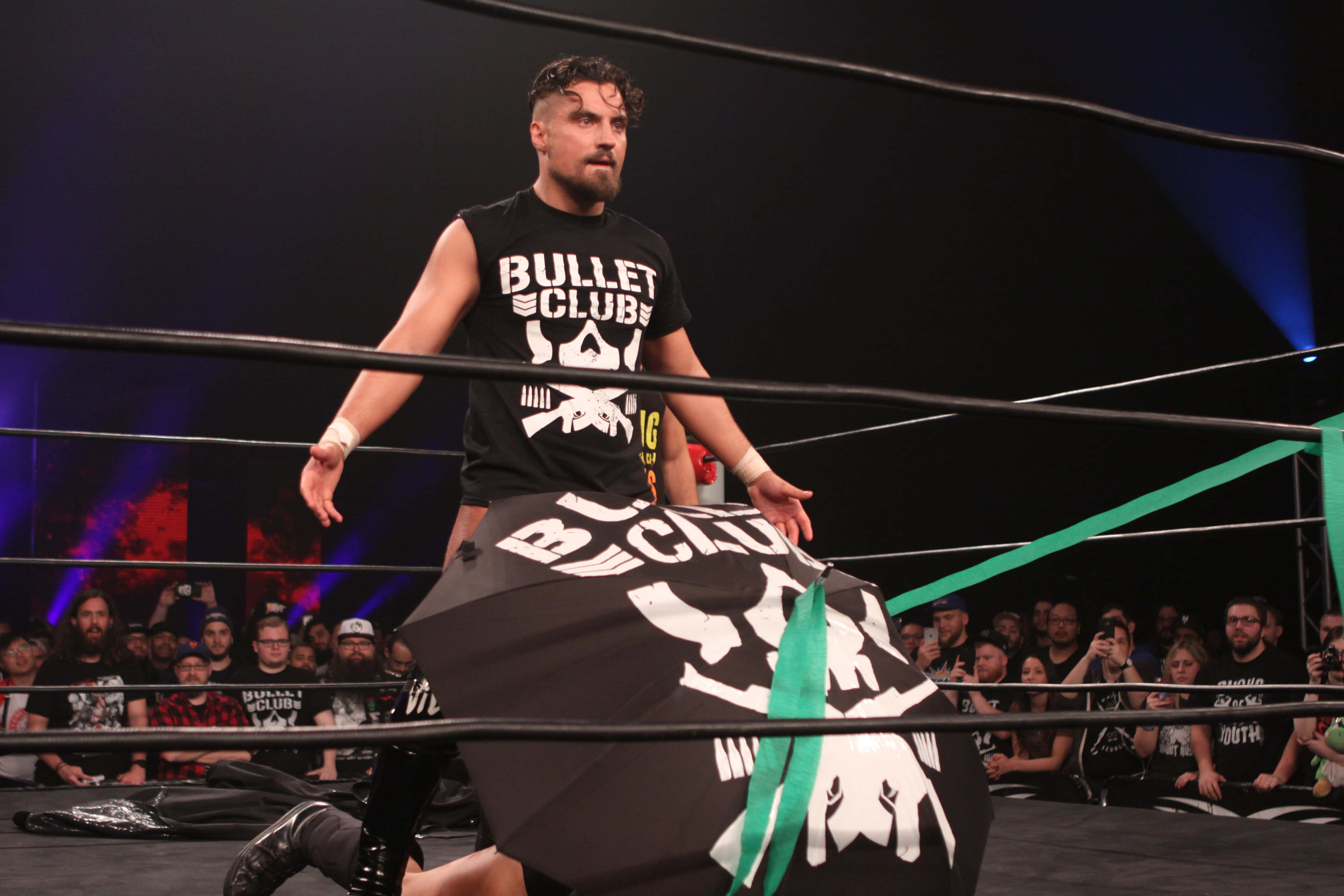 English 桌布 壁紙 — 一名身穿黑色 'Bullet Club' T恤的摔角手站在擂台上，手持相配的旗幟，粉絲們在舞台燈光下觀看。