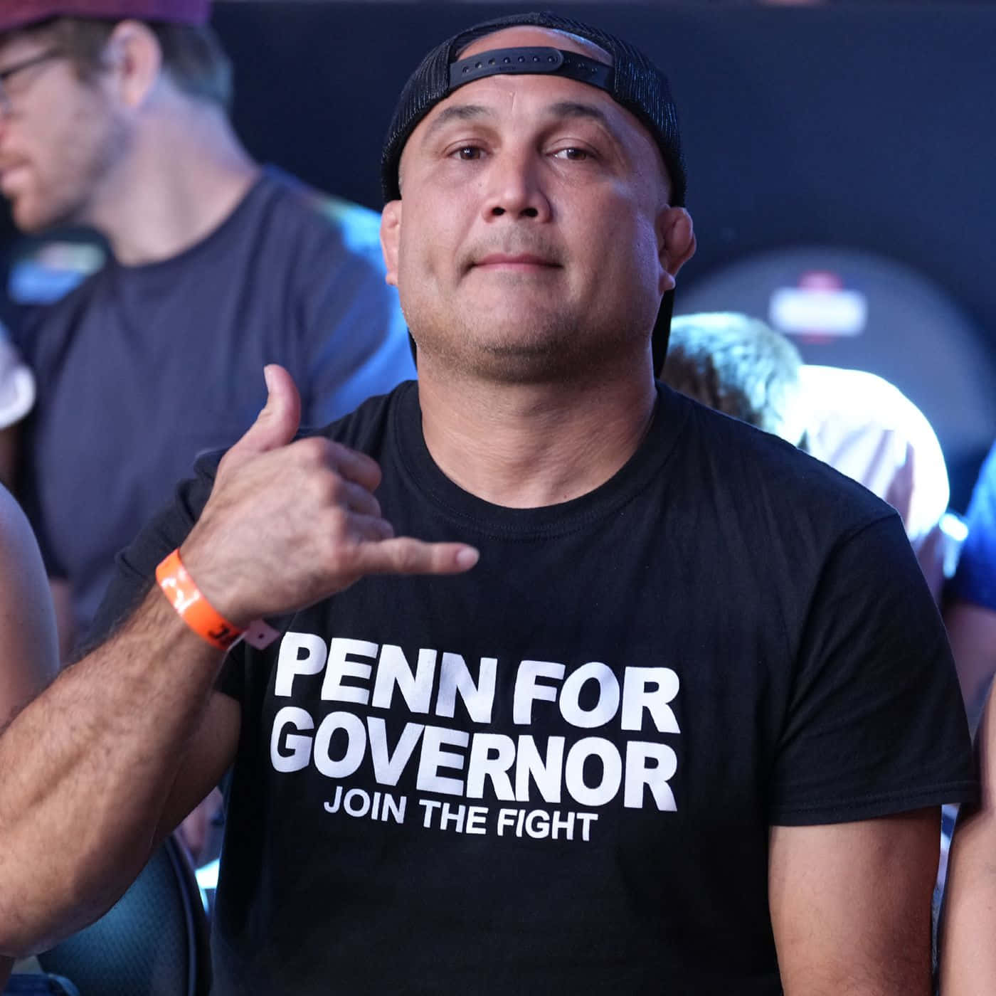 MMA 桌布 壁紙 — 一名身穿黑色「PENN FOR GOVERNOR」T恤的男子在活動中做出沙卡手勢，身後有模糊的與會者。