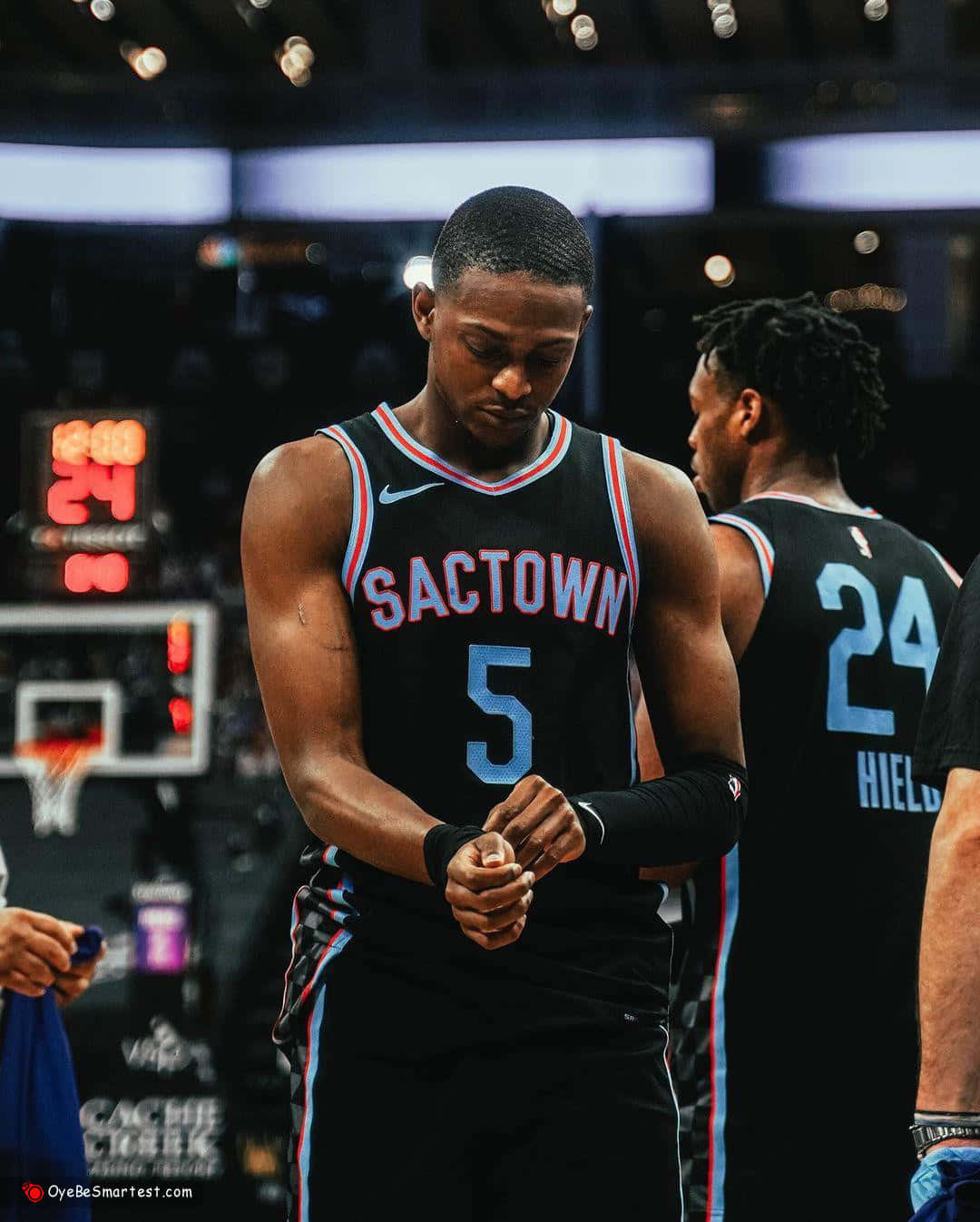 De'aaron Fox 桌布 壁紙 — 一名身穿黑色 'SACTOWN' 球衣、背號5的籃球運動員在場館燈光下低頭調整護腕。