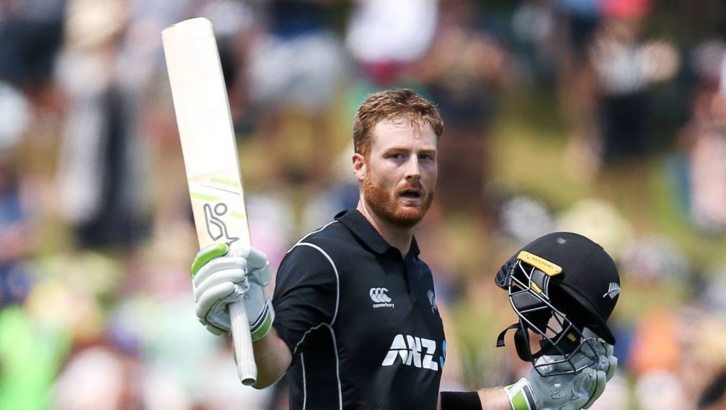 martin guptill 桌布 壁紙 — 一名身穿黑色ANZ球衣的板球運動員，手持球棒和頭盔，站在陽光明媚的球場上，背景是模糊的觀眾。