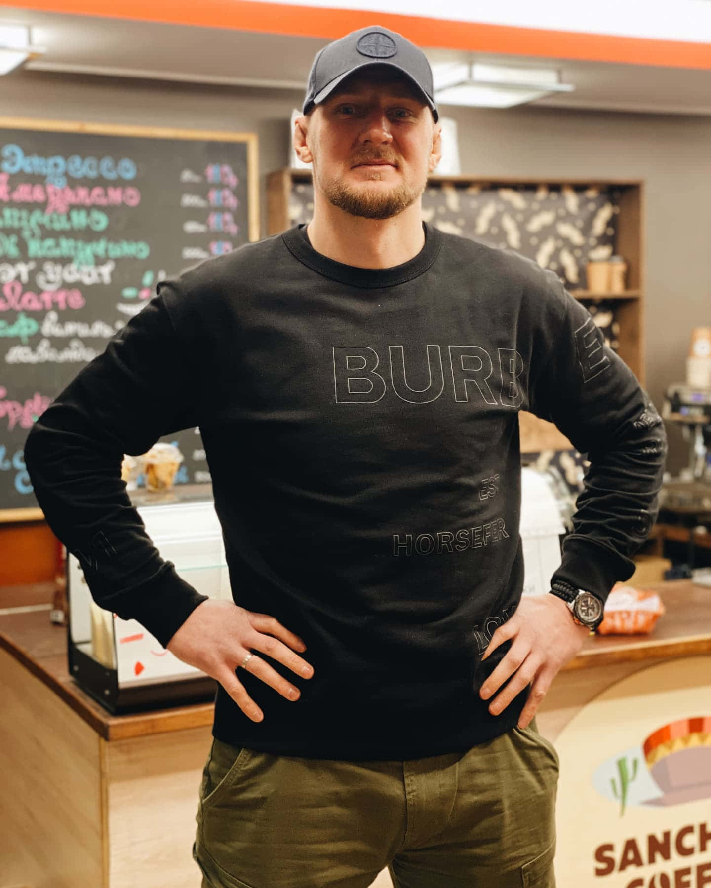 Celebrity 桌布 壁紙 — 一名身穿黑色Burberry衛衣和帽子的男子，在舒適的咖啡店內自信地雙手叉腰站立。