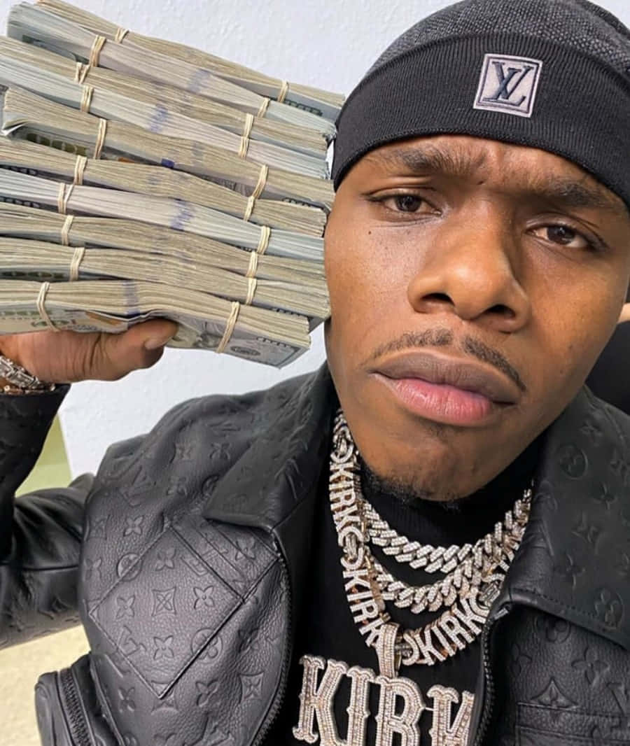 Dababy 桌布 壁紙 — 一名身穿黑色Louis Vuitton外套和毛帽的男子，手持一大疊捆好的現金，專注地凝視鏡頭。
