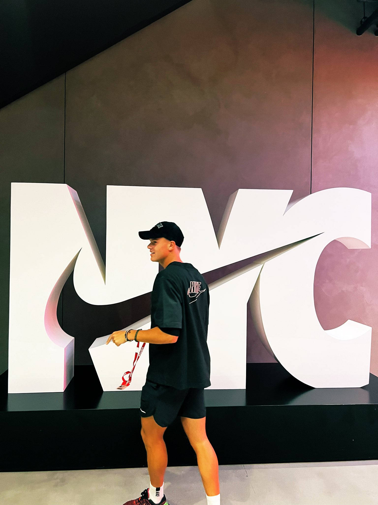 Champion 桌布 壁紙 — 一名身穿黑色Nike裝備的年輕男子站在一個帶有勾號的巨型白色NYC標誌旁，背景是漸層牆。