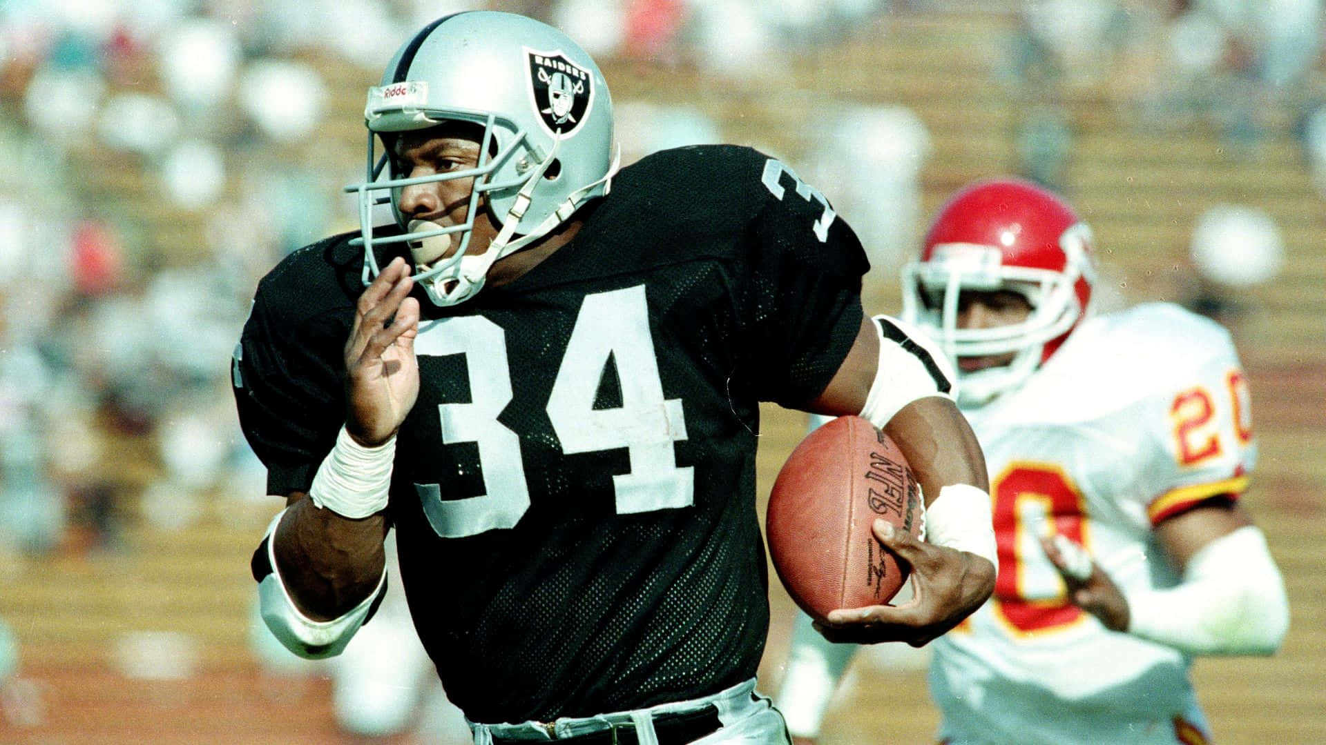 Bo Jackson 桌布 壁紙 — 一名身穿黑色Raiders球衣、號碼34的美式足球員在陽光普照的球場上帶球奔跑，一名身穿紅色的防守球員在後面追趕。