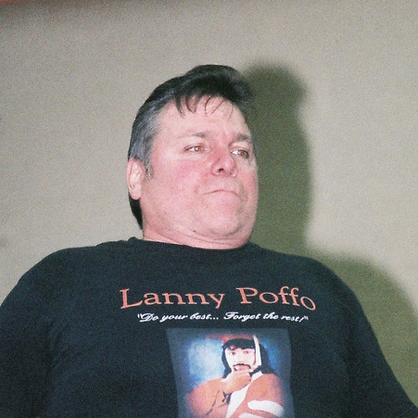 摔角 桌布 壁紙 — 一名身穿黑色T恤、印有Lanny Poffo及「do your best forget the rest」字句的男子，靠著素色牆壁站立。