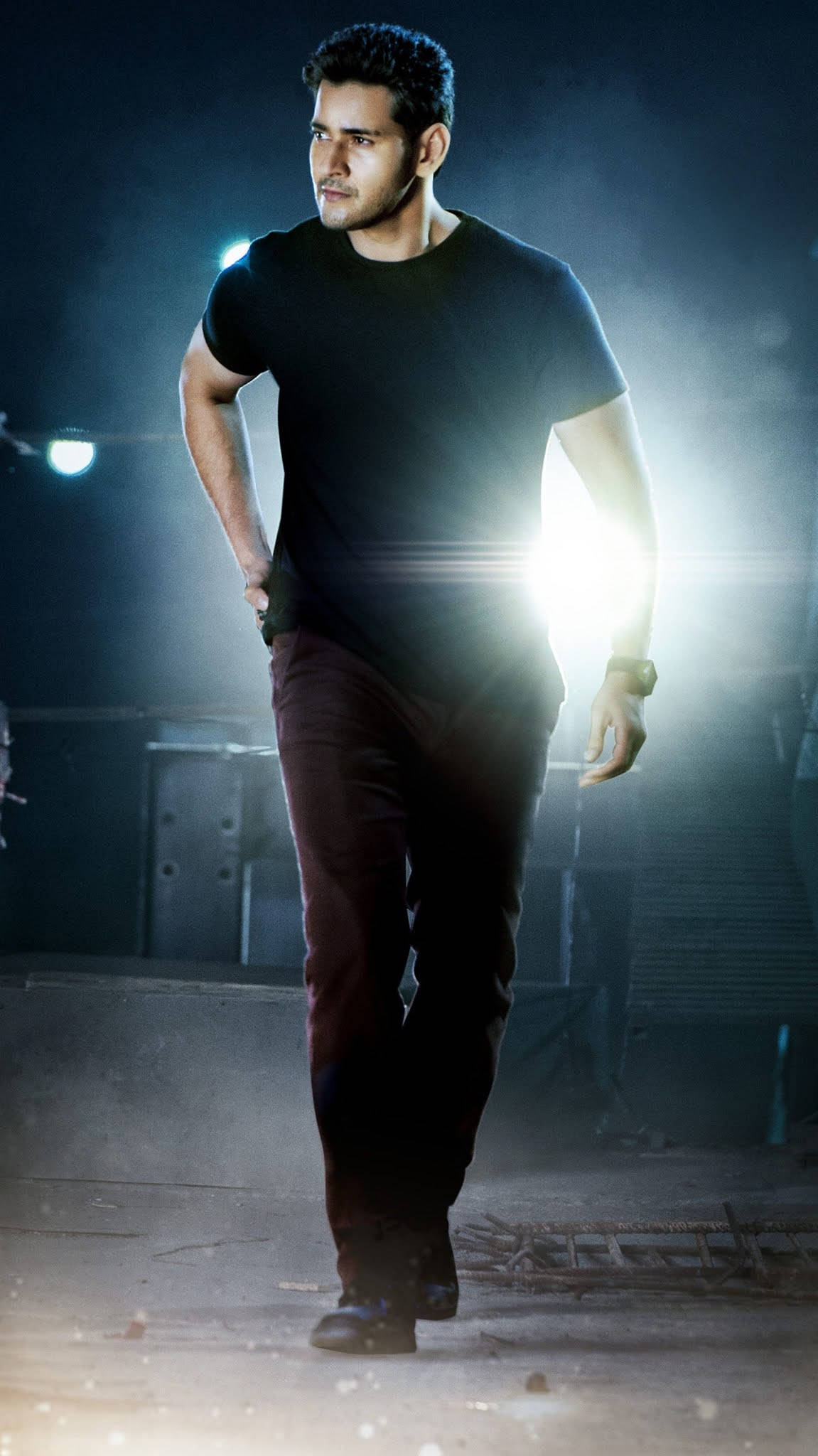 Mahesh Babu HD 桌布 壁紙 — 一名身穿黑色T恤和褲子的男子在昏暗的工業環境中，在戲劇性的舞台燈光下大步向前走。