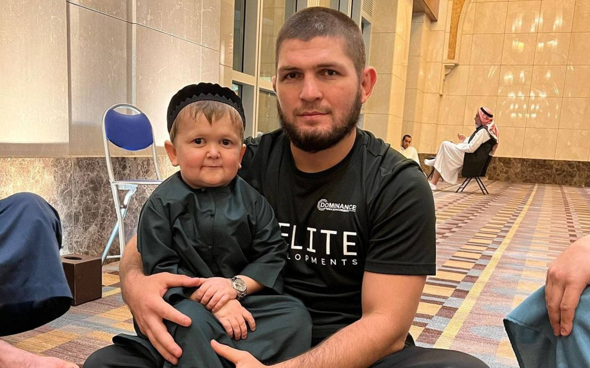 khabib 桌布 壁紙 — 一名身穿黑色T恤的男子坐在寬敞的大理石地板大廳中，抱著一名穿著傳統服飾的小男孩。