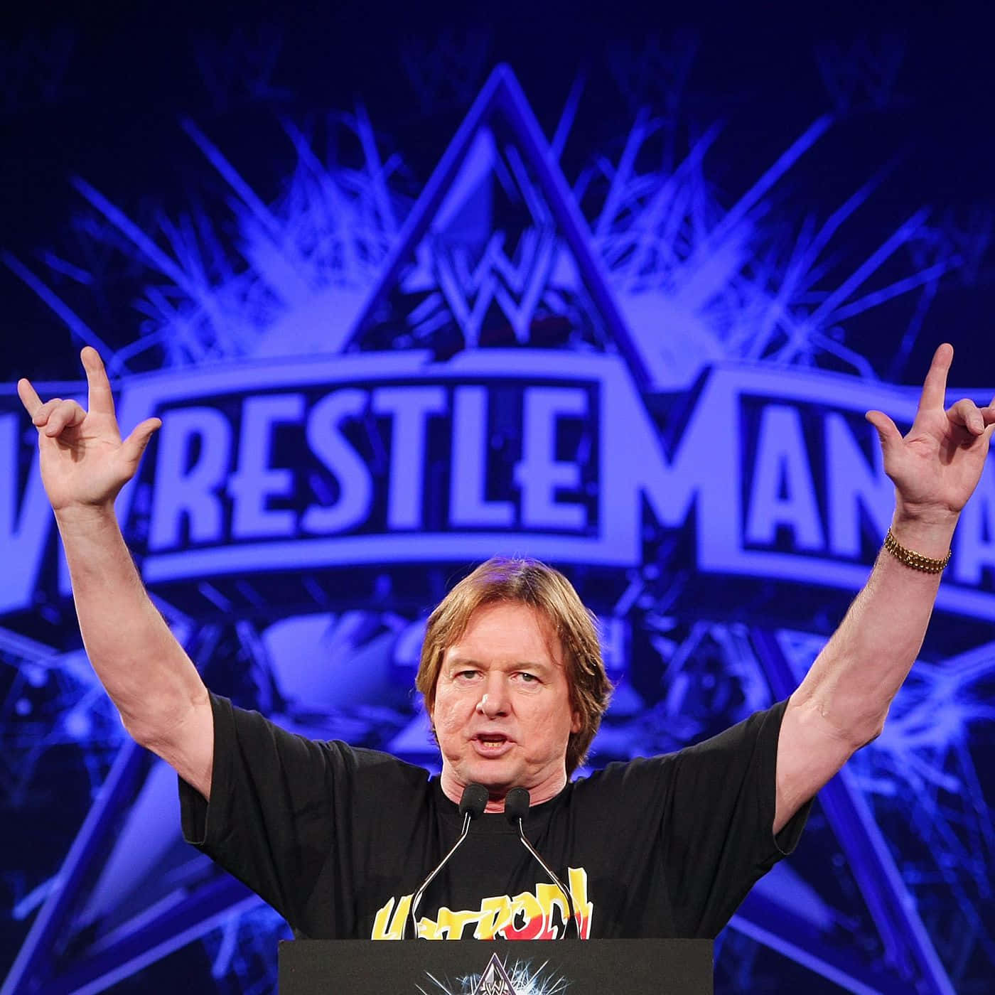 摔角 桌布 壁紙 — 一名身穿黑色T恤的男子站在講台上，在明亮的藍色WrestleMania舞台燈光下做出搖滾手勢。