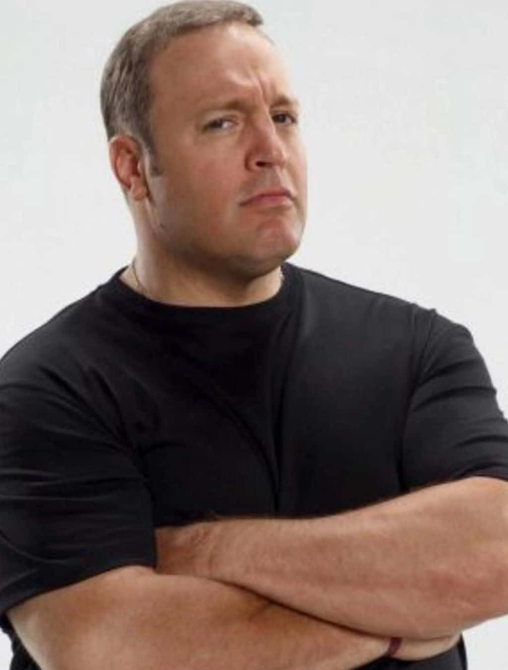 Kevin James 桌布 壁紙 — 一名身穿黑色T恤的男子雙臂交叉，站在純白色背景前，表情嚴肅地望向一旁。