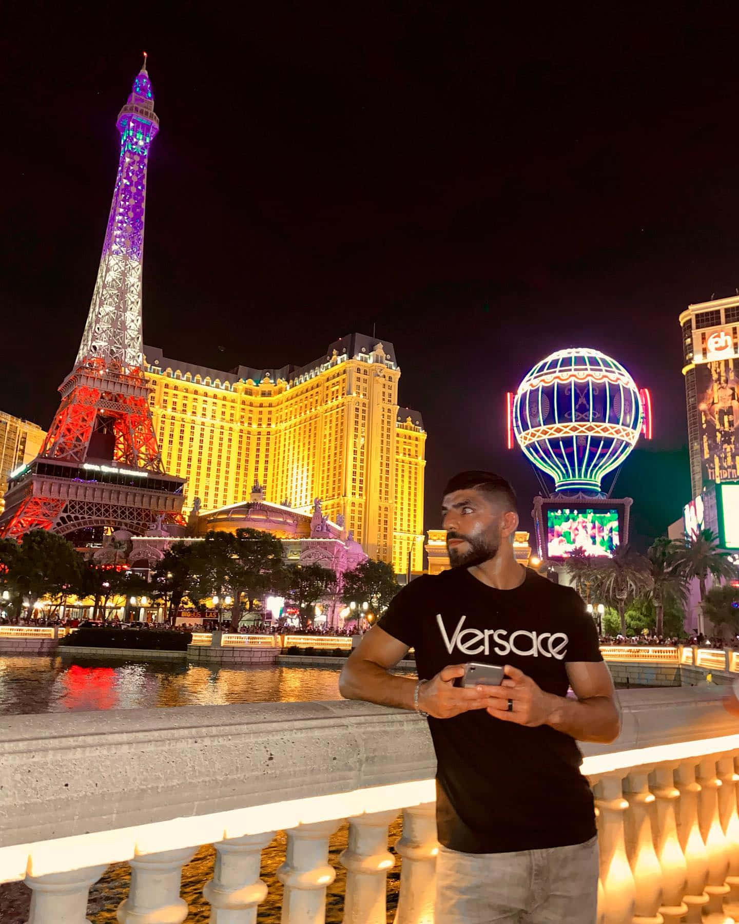 拉斯維加斯 桌布 壁紙 — 一名身穿黑色Versace T恤的男子在夜晚站在發光的欄杆旁，凝視著拉斯維加斯耀眼的複製艾菲爾鐵塔和明亮的賭場燈光。
