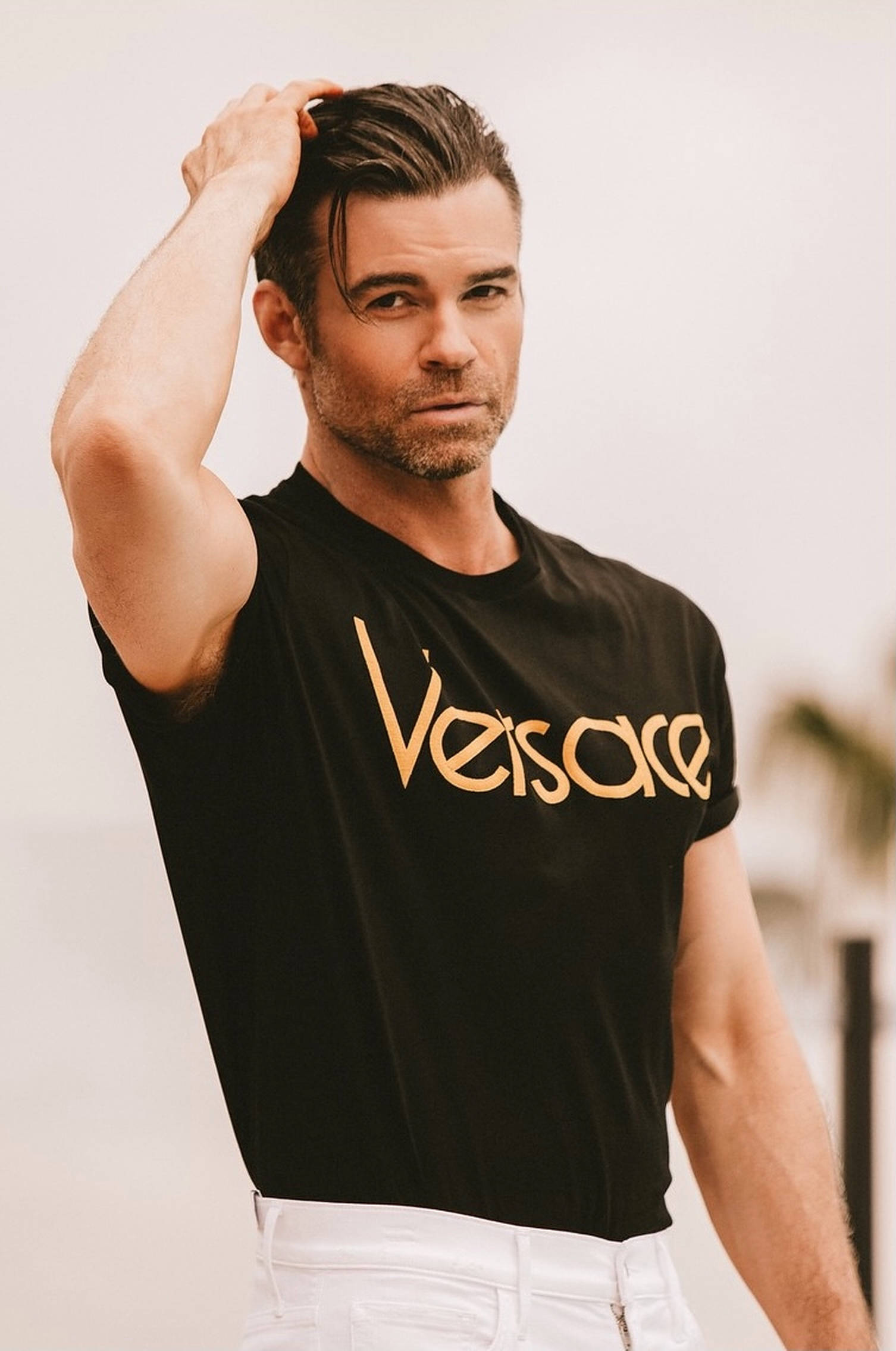Daniel Gillies 桌布 壁紙 — 一名身穿黑色Versace T恤和白色褲子的男子，在柔和模糊的背景前，用手梳理他造型過的頭髮。