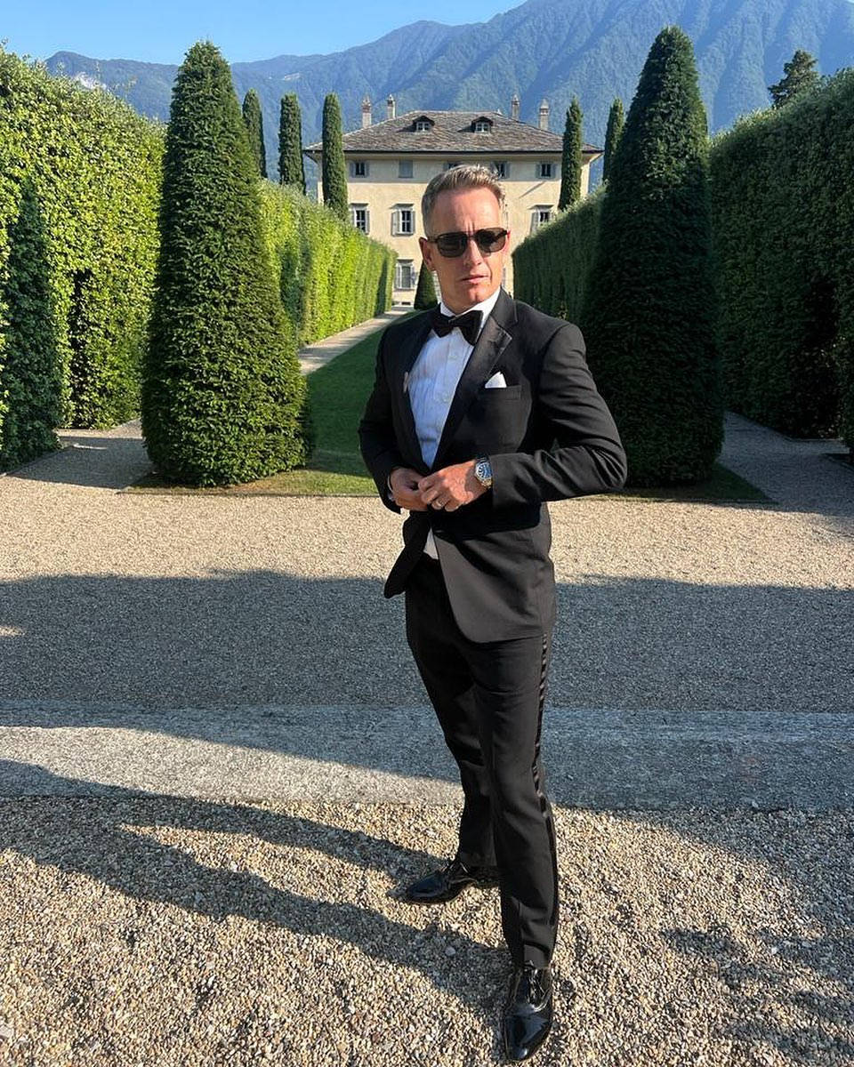 Tuxedo 桌布 壁紙 — 一名身穿黑色修身燕尾服和太陽眼鏡的男子自信地站在一條碎石小徑上，小徑兩旁是修剪整齊的樹籬，背景有山脈和一座別墅。