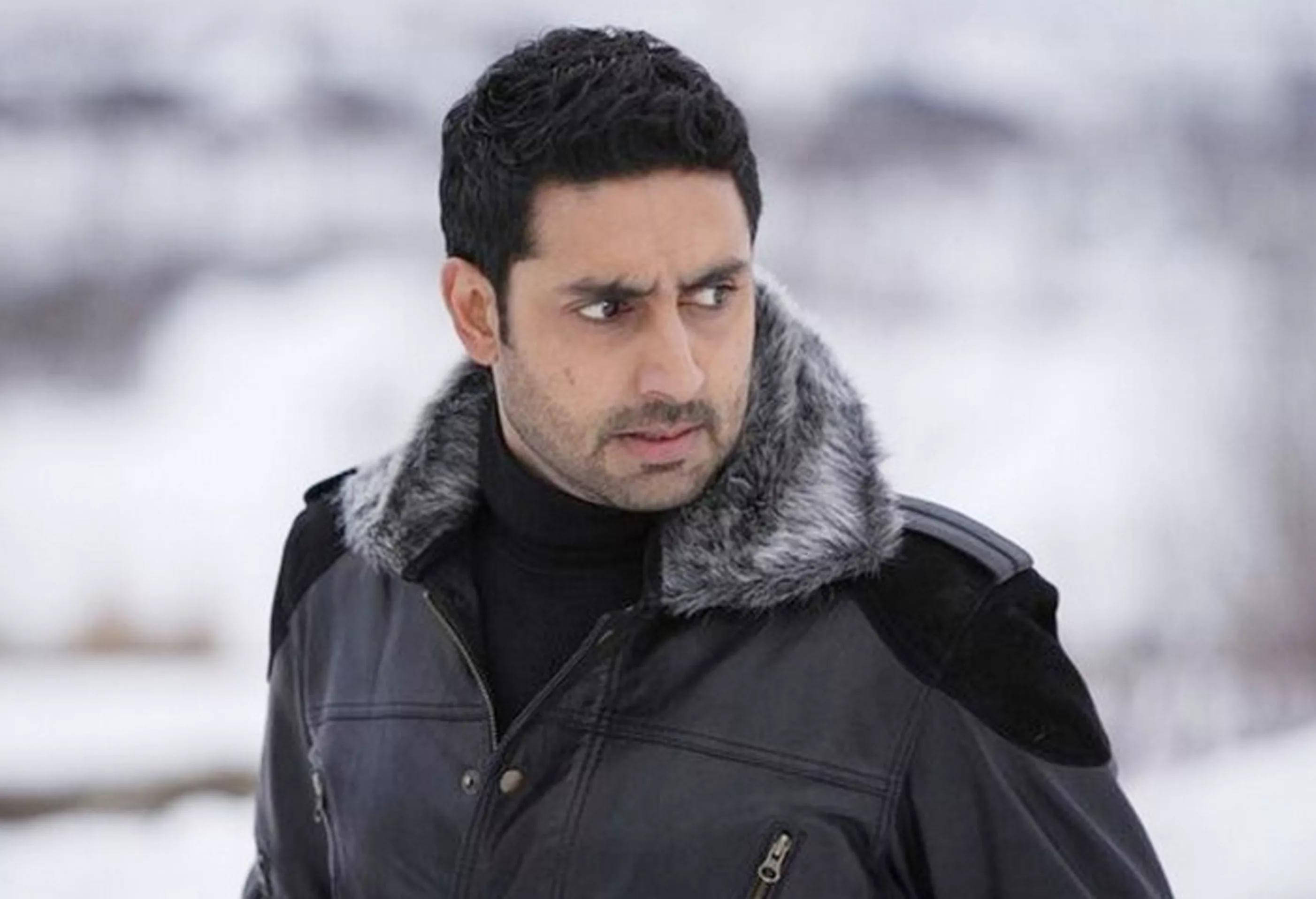 abhishek bachchan 桌布 壁紙 — 一名身穿黑色冬季外套、帶有毛皮襯裡連帽的男子站在雪景中，表情嚴肅地望向一旁。
