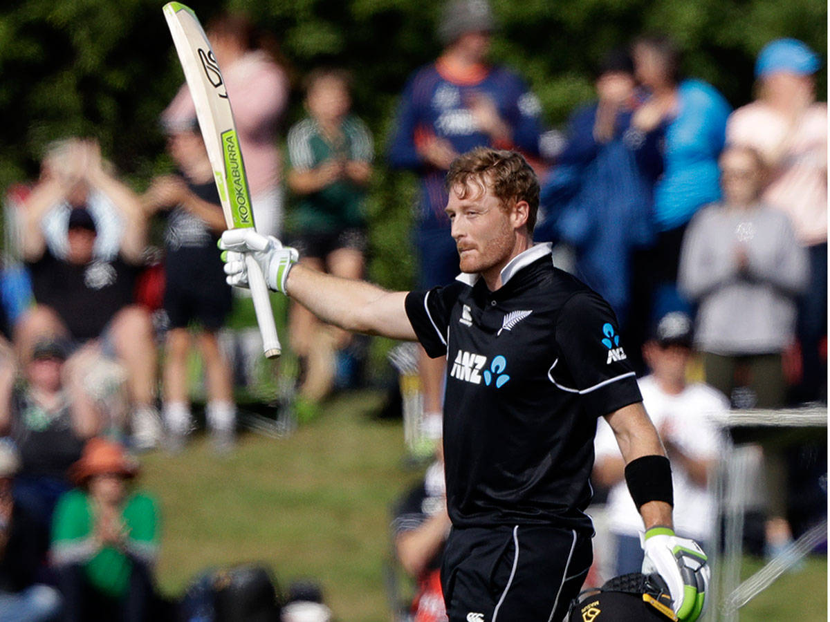 martin guptill 桌布 壁紙 — 一名身穿黑色制服的新西蘭板球運動員在陽光明媚的球場上高舉球棒，球迷們鼓掌歡呼。