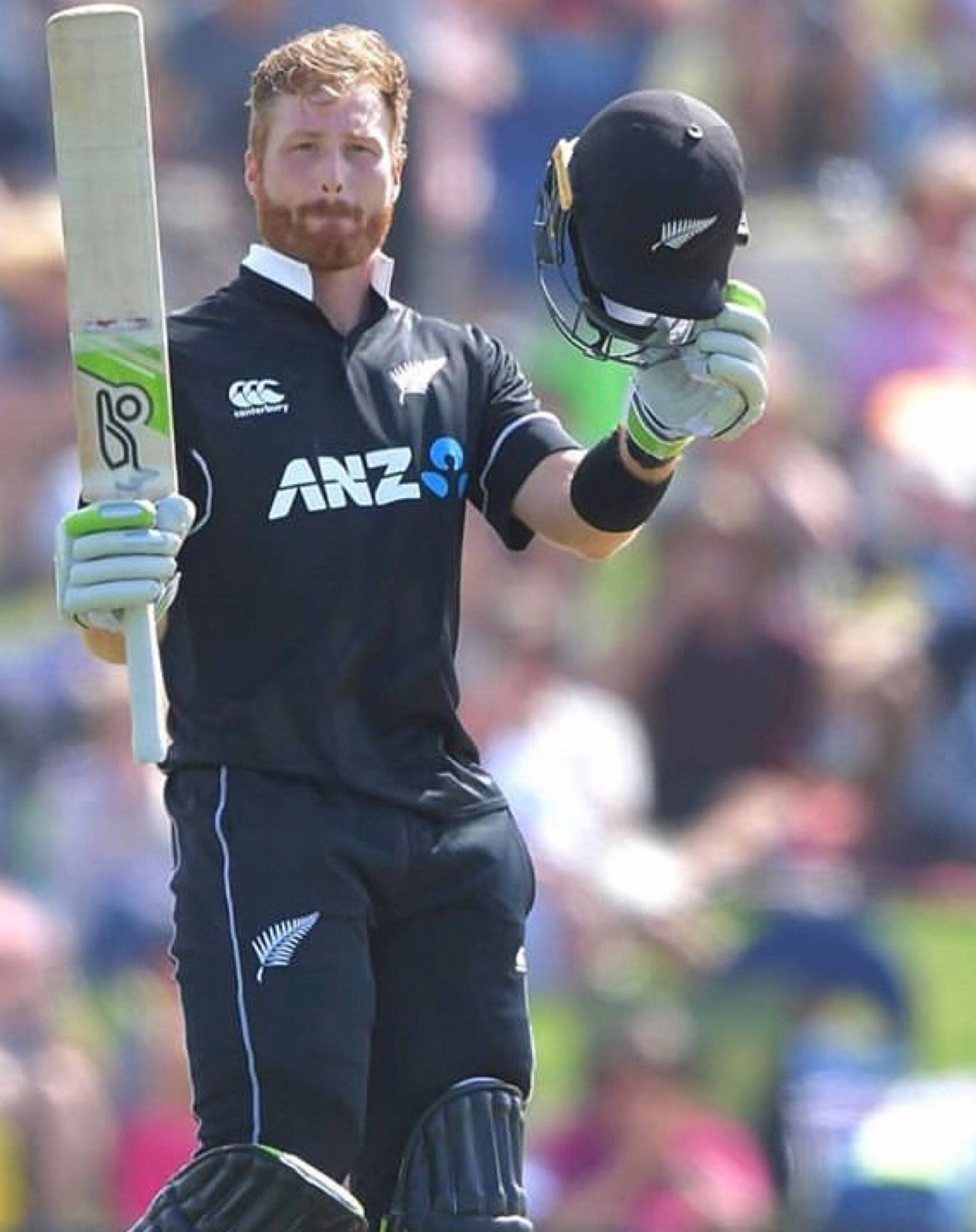 martin guptill 桌布 壁紙 — 一名身穿黑色制服的新西蘭板球運動員在明亮的日光下手持球棒和頭盔，向歡呼的人群致意。