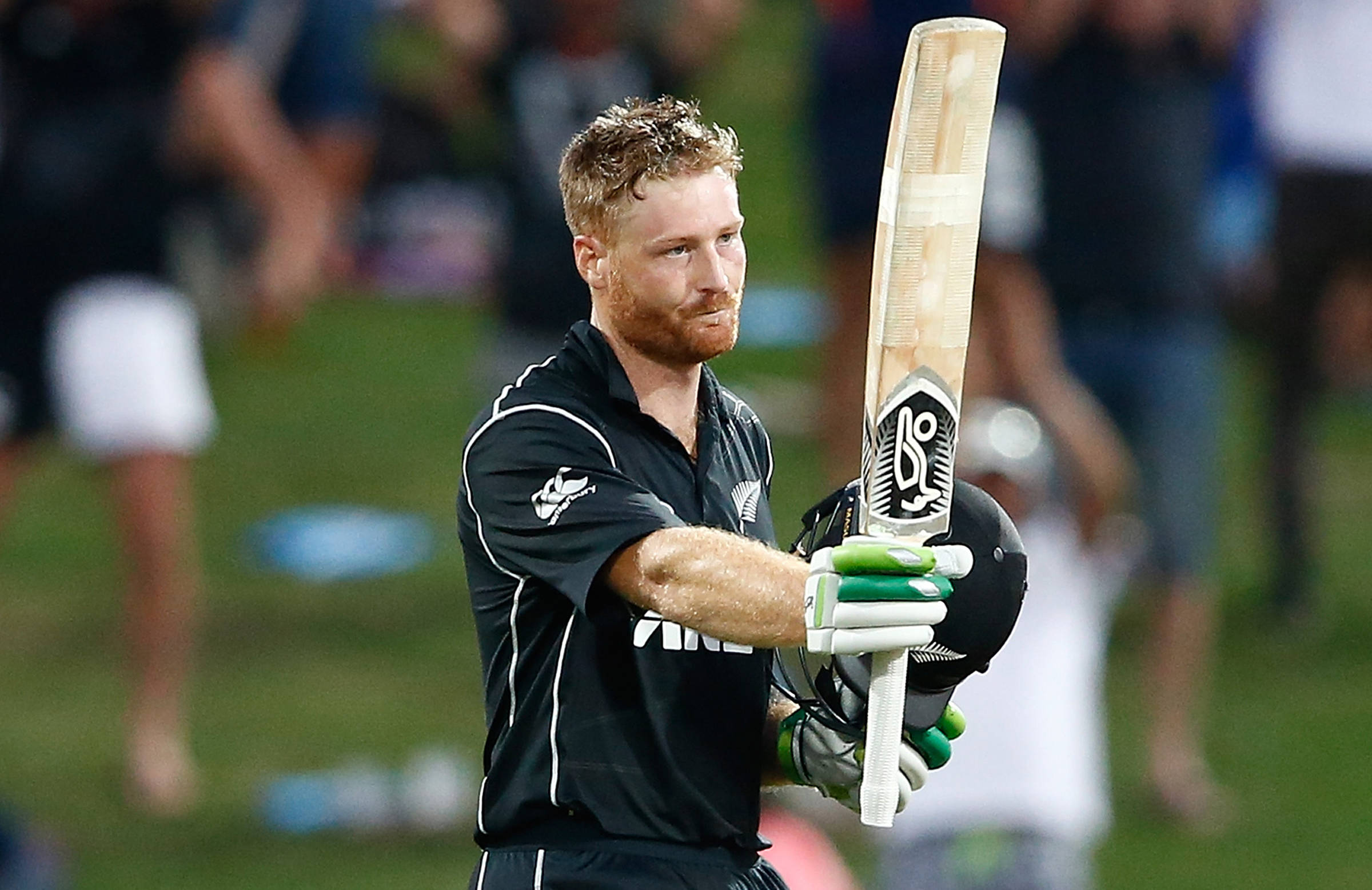 martin guptill 桌布 壁紙 — 一名身穿黑色制服的板球選手在球場燈光下，於綠色場地上高舉球棒，手持頭盔。