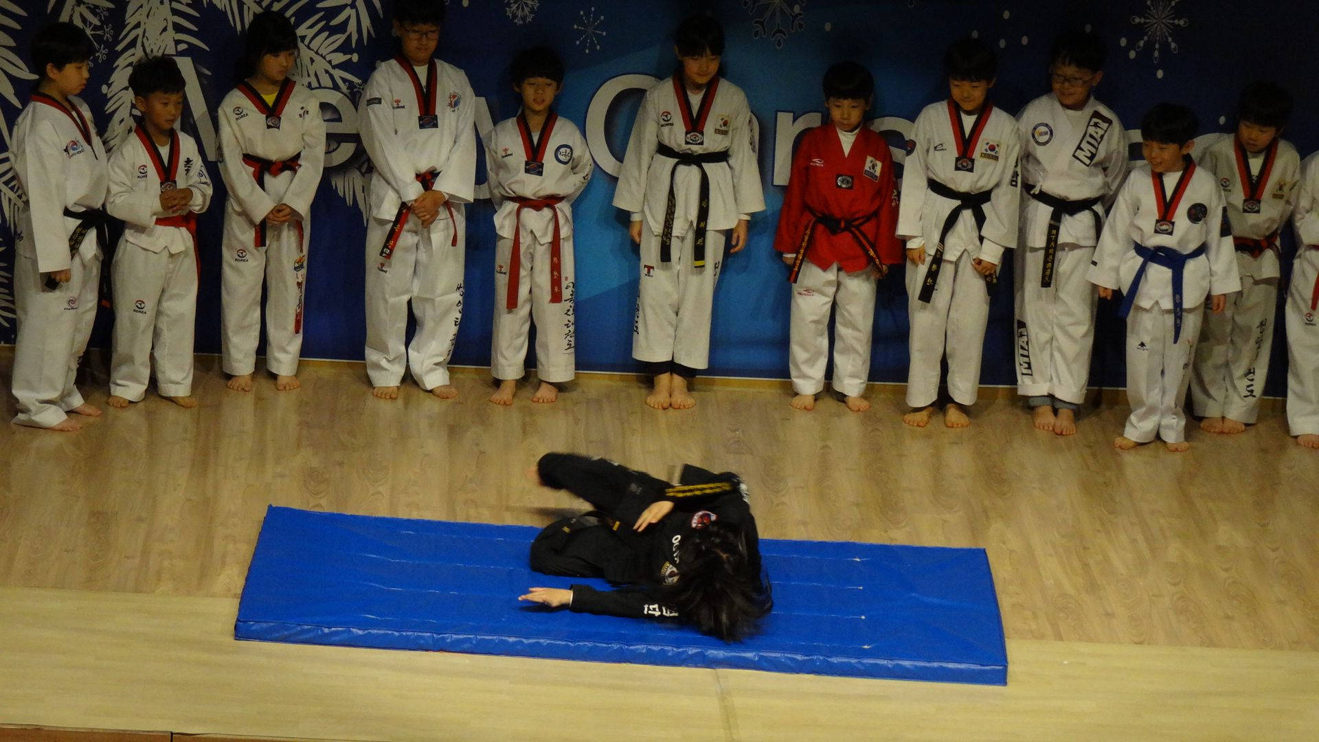 MMA 桌布 壁紙 — 一名身穿黑色制服的武術家在藍色墊子上表演地板動作，而一排身穿白色制服的年輕學生則專注地觀看。