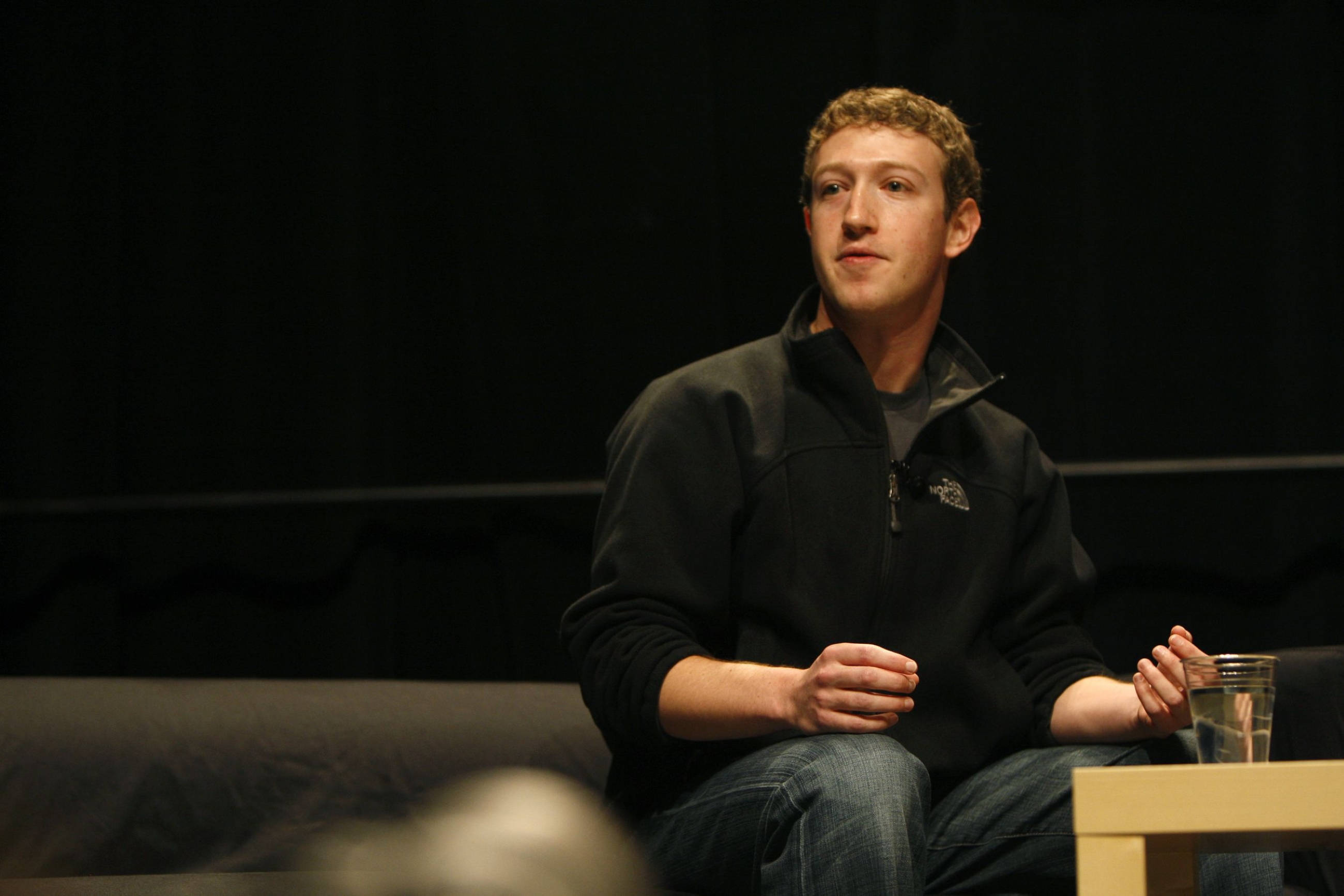 Zuckerberg 桌布 壁紙 — 一名身穿黑色刷毛外套的男子坐在沙發上，背景昏暗，旁邊的小桌上放著一個玻璃杯。