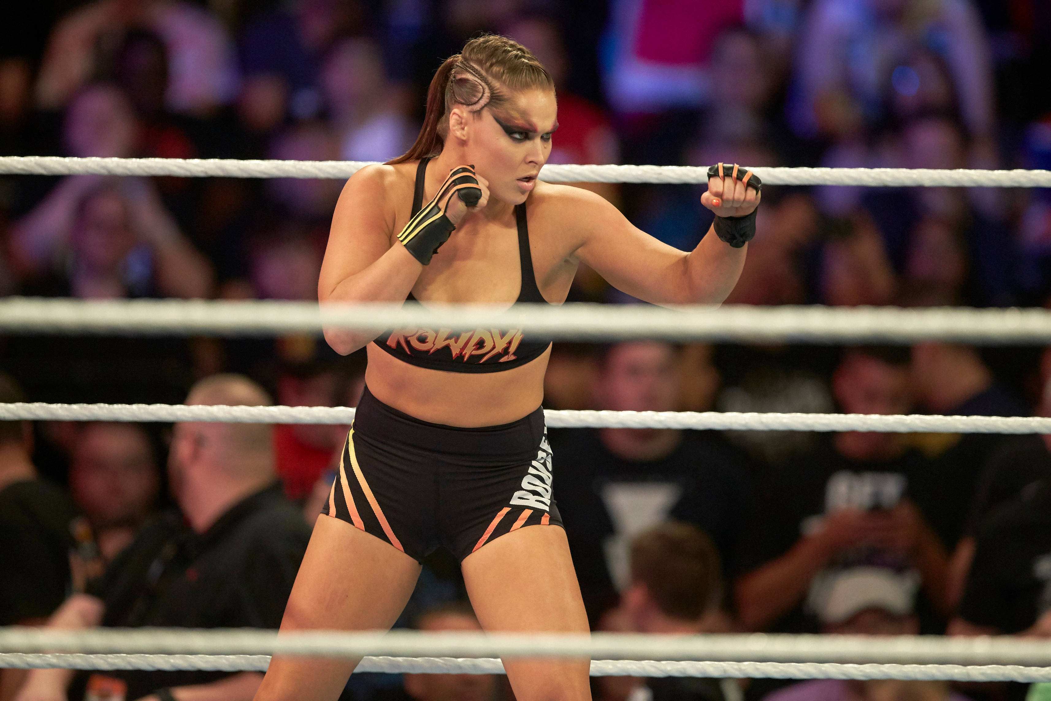 Rousey 桌布 壁紙 — 一名身穿黑色和橙色裝備的女摔角手在摔角台上舉起拳頭，人群在繩索後模糊不清。