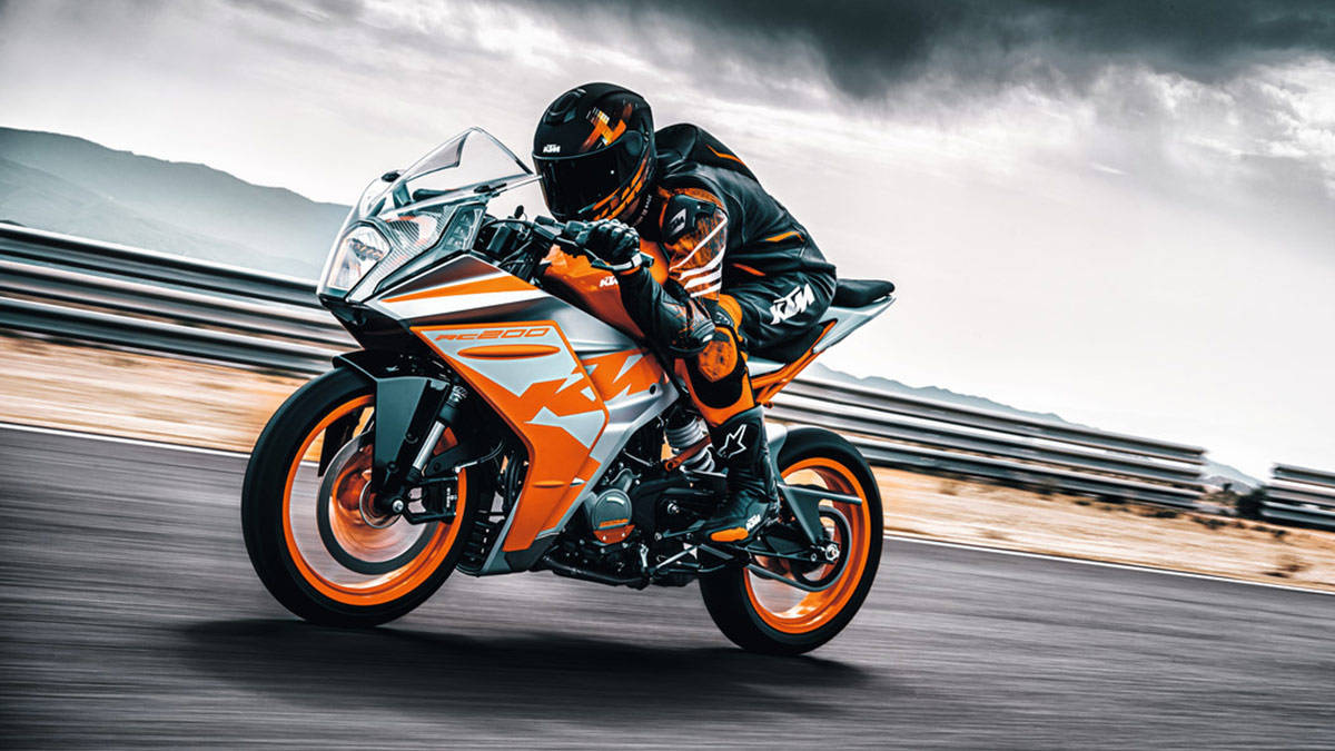 KTM RC 200 桌布 壁紙 — 一名身穿黑色和橙色裝備的騎士在暴風雨天空下的賽道上，騎著鮮豔的橙色和銀色KTM機車傾斜入彎。