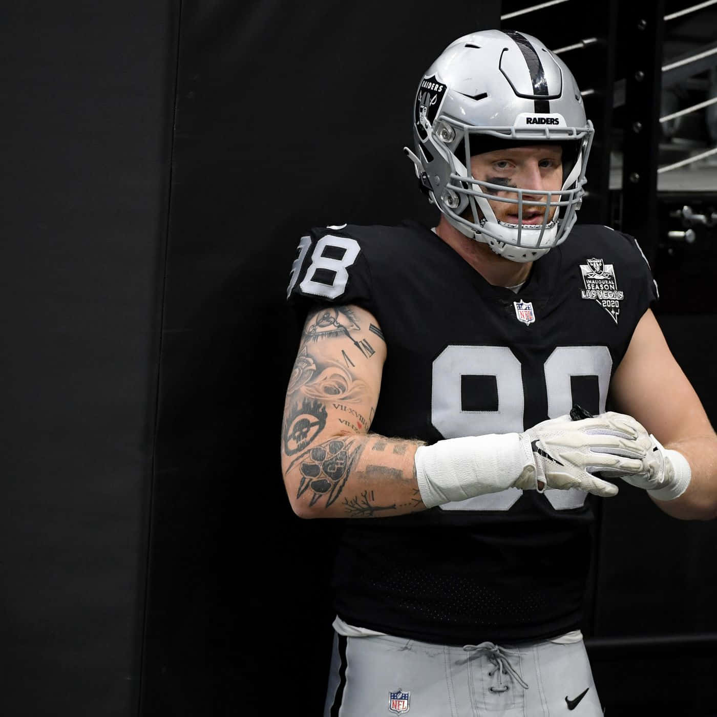運動 桌布 壁紙 — 一名身穿黑色和銀色 Raiders 球衣的足球員站在燈光昏暗的更衣室裡，手臂上的刺青清晰可見。