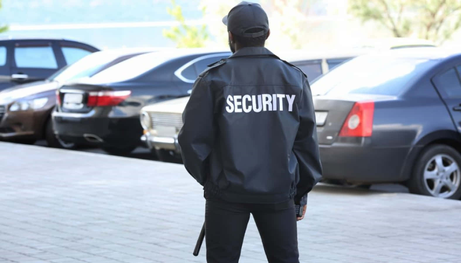 security 桌布 壁紙 — 一名身穿黑色外套、背後印有「SECURITY」字樣的保全人員，在鋪砌的停車場中警覺地站立，周圍是汽車。