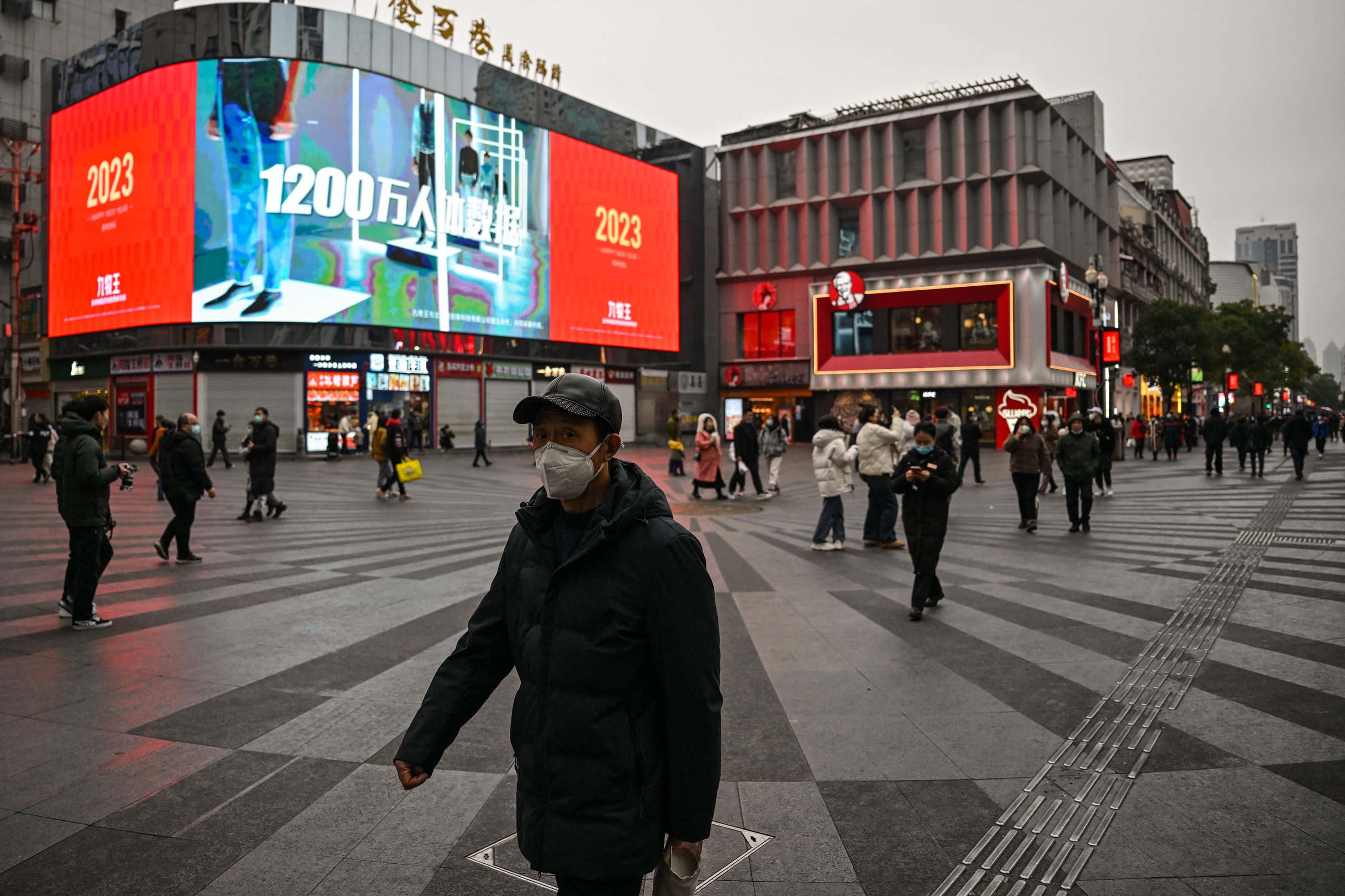 中國 桌布 壁紙 — 一名身穿黑色外套和口罩的男子，在陰天之下走過寬闊的城市廣場，廣場上有發光的紅色數位廣告看板顯示2023年。