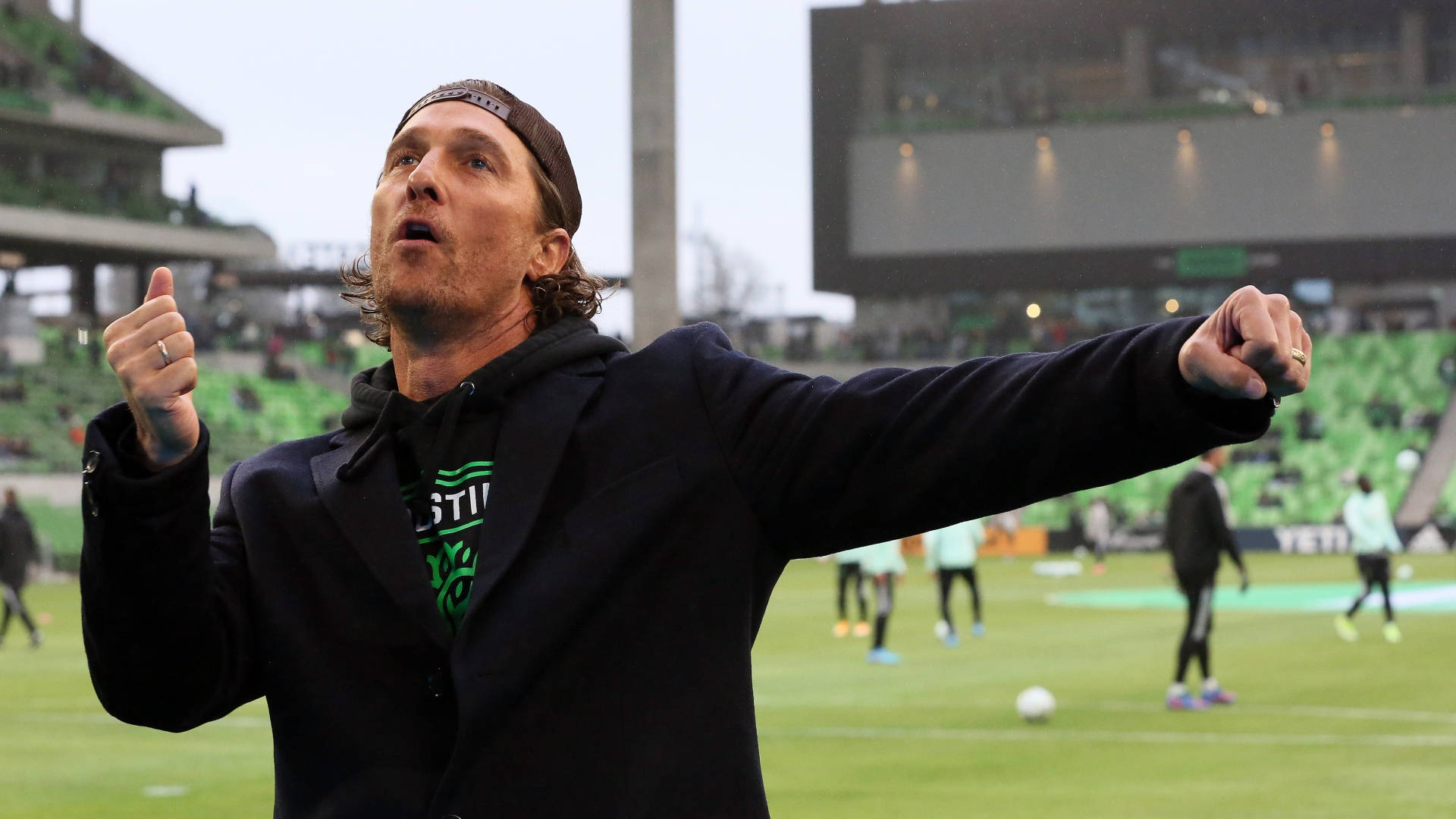 Austin Fc 桌布 壁紙 — 一名身穿黑色外套和帽子的男子在綠色足球場上熱情慶祝，背景是體育場看台。