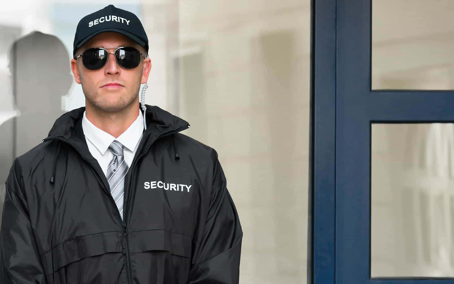 security 桌布 壁紙 — 一名身穿黑色外套和帽子的警衛，神情嚴肅地警覺地站在玻璃門附近。